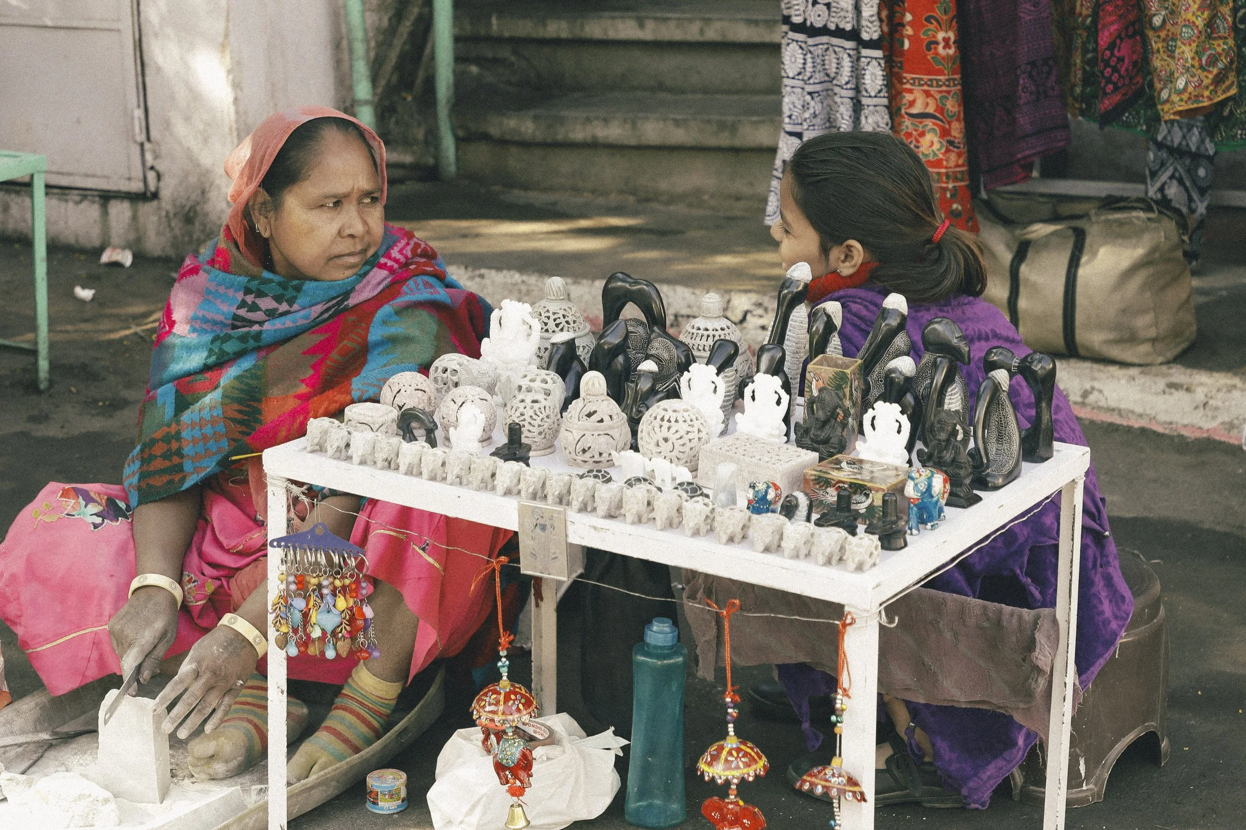 A woman and a girl sitting across from each other at a street market stall with a table displaying carved jade or stone ornaments, small statues, and decorative items, with colorful textiles hanging in the background.