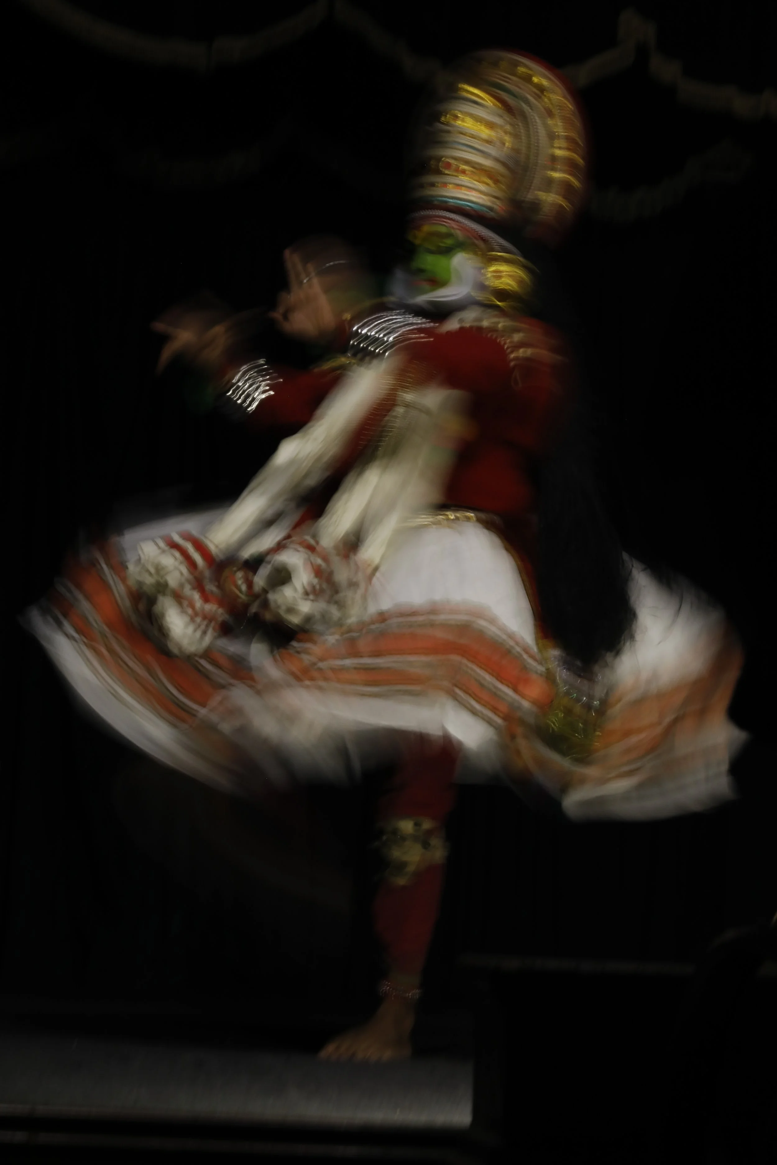 A dancer performing a traditional Indian dance, dressed in colorful attire with elaborate jewelry and a decorated headpiece, captured in motion against a black background.