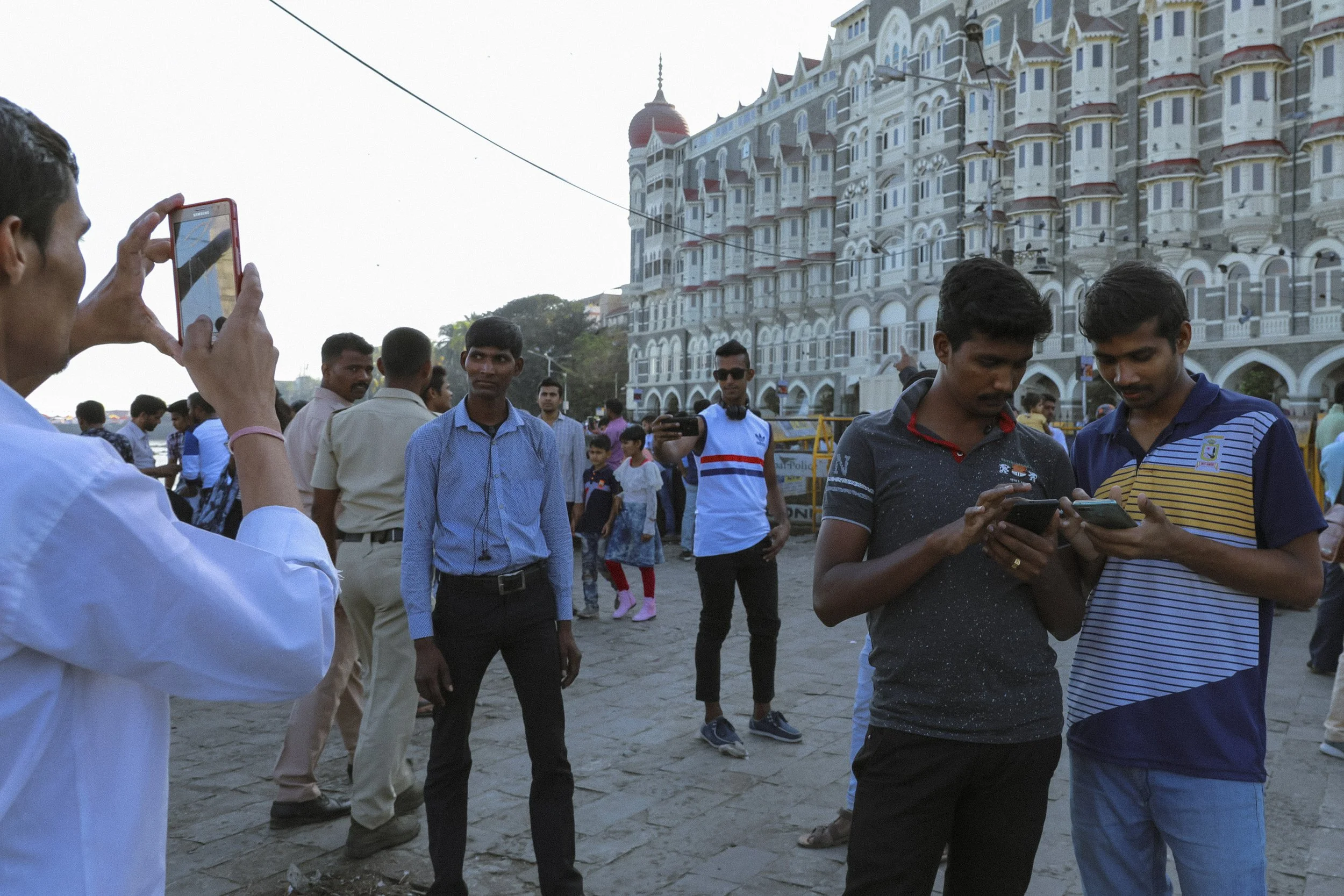 A group of people, mostly young men, are gathered outside a large ornate building, taking pictures and looking at their phones during the daytime.
