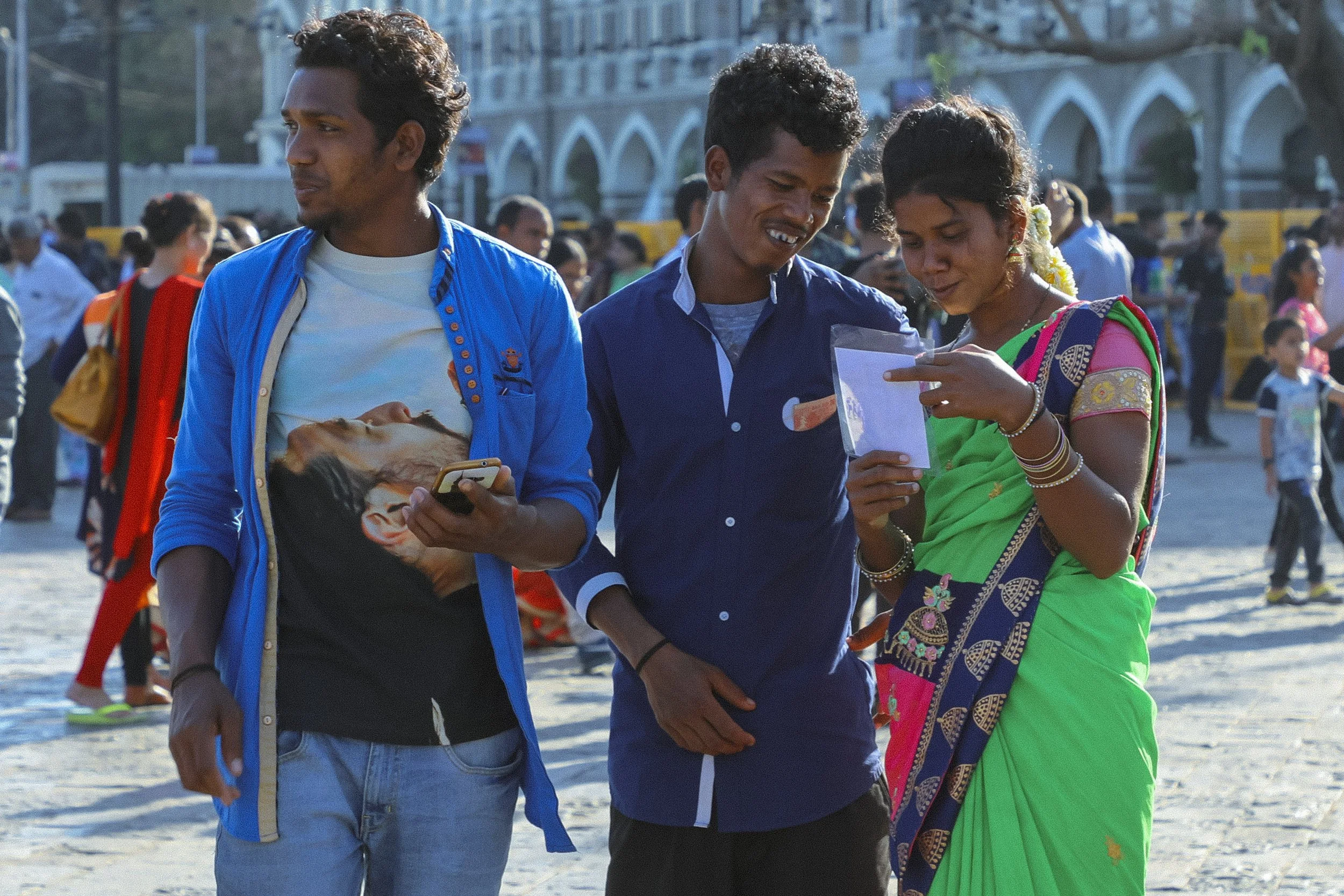 Three people standing outdoors in a busy area, looking at a piece of paper or photograph together. The man on the left is wearing a blue jacket over a t-shirt, the man in the middle is dressed in a blue shirt, and the woman on the right is in a tradi