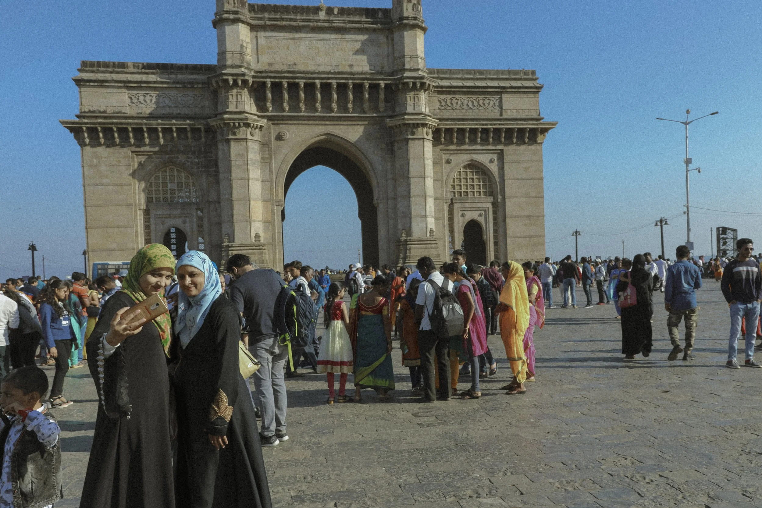 Crowds of people gathered near the India Gate monument, with many women wearing colorful traditional Indian clothing. The weather is sunny with clear blue skies.