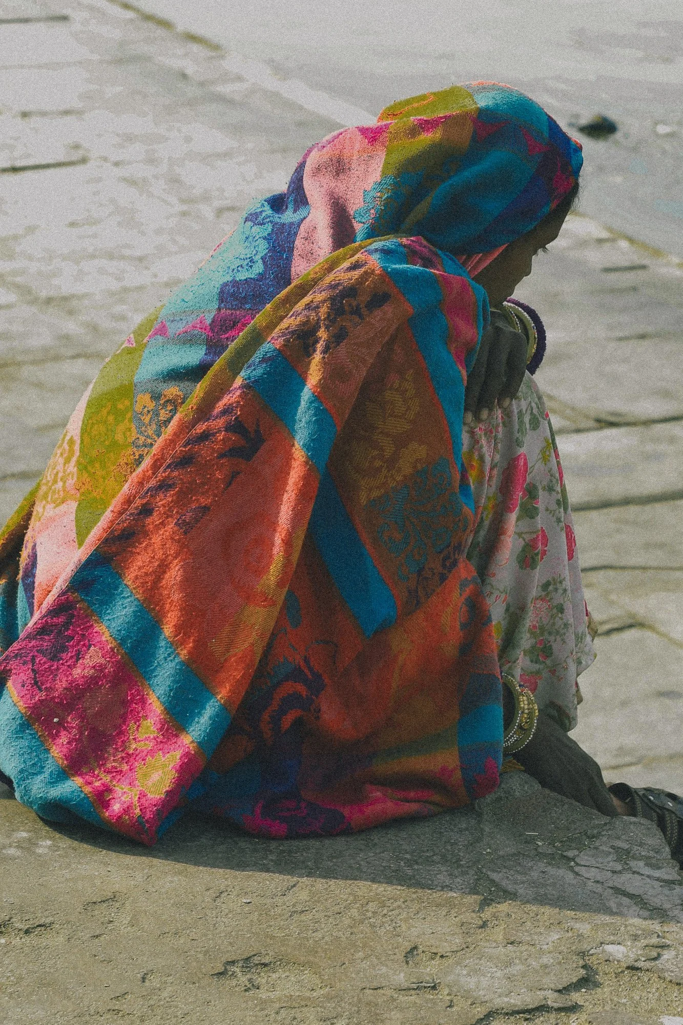 A woman wearing a colorful patterned headscarf and dress sitting on a stone surface by water, with her head bowed down.