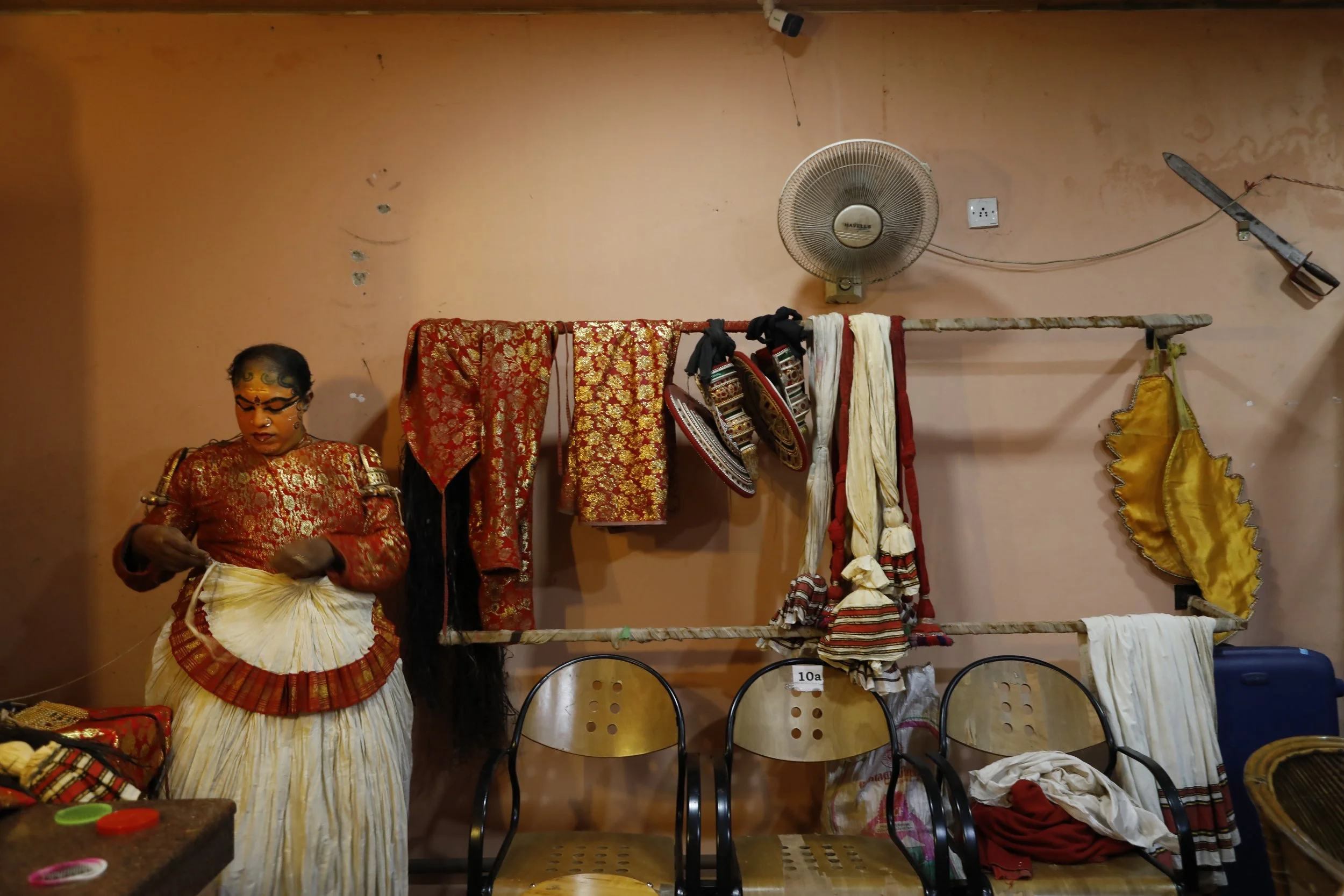 A person dressed in traditional Indian attire is standing next to a display of fabric and accessories hanging on a wall, with chairs in front.