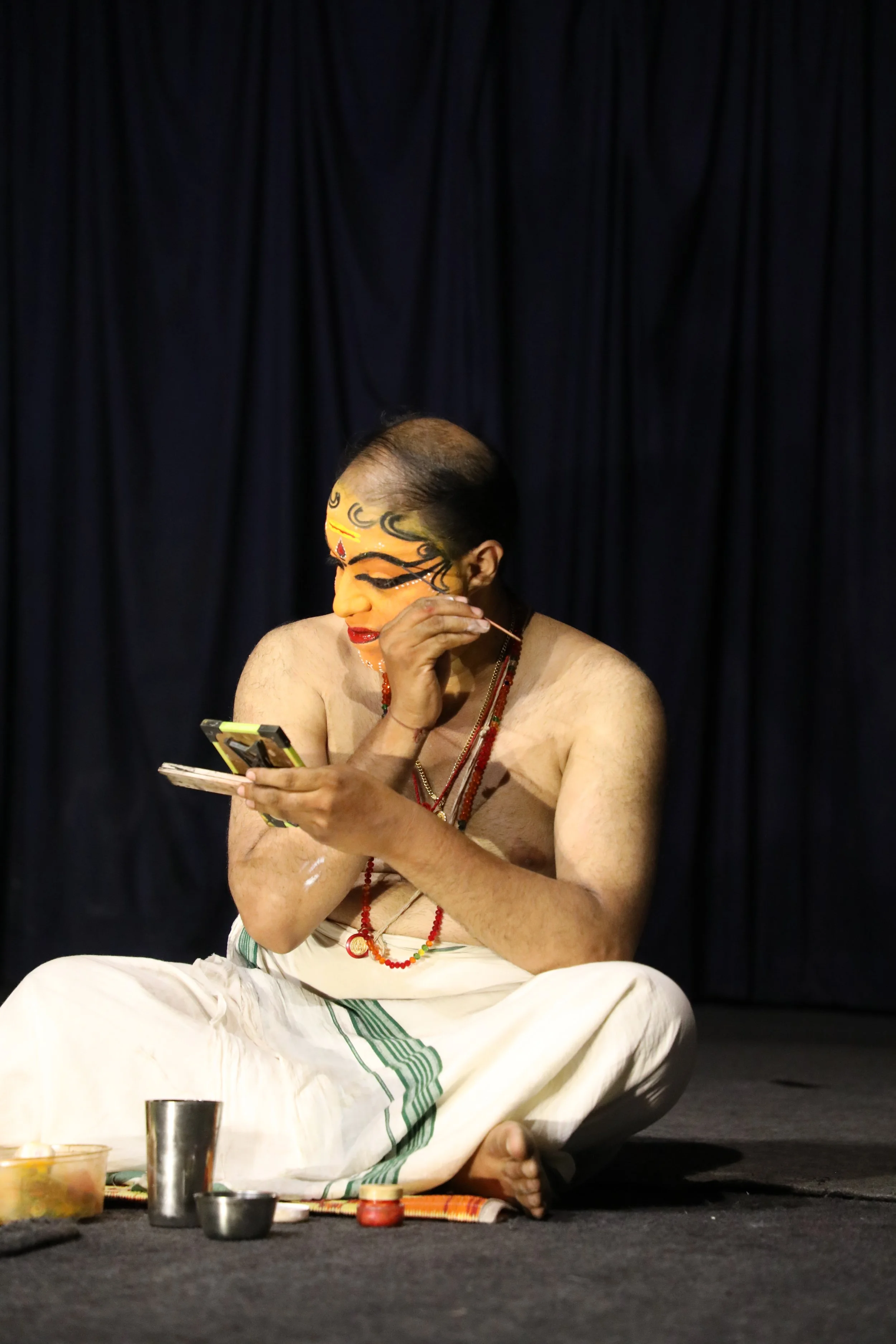 A person dressed in traditional Indian attire with makeup, sitting cross-legged on stage, looking at a phone.