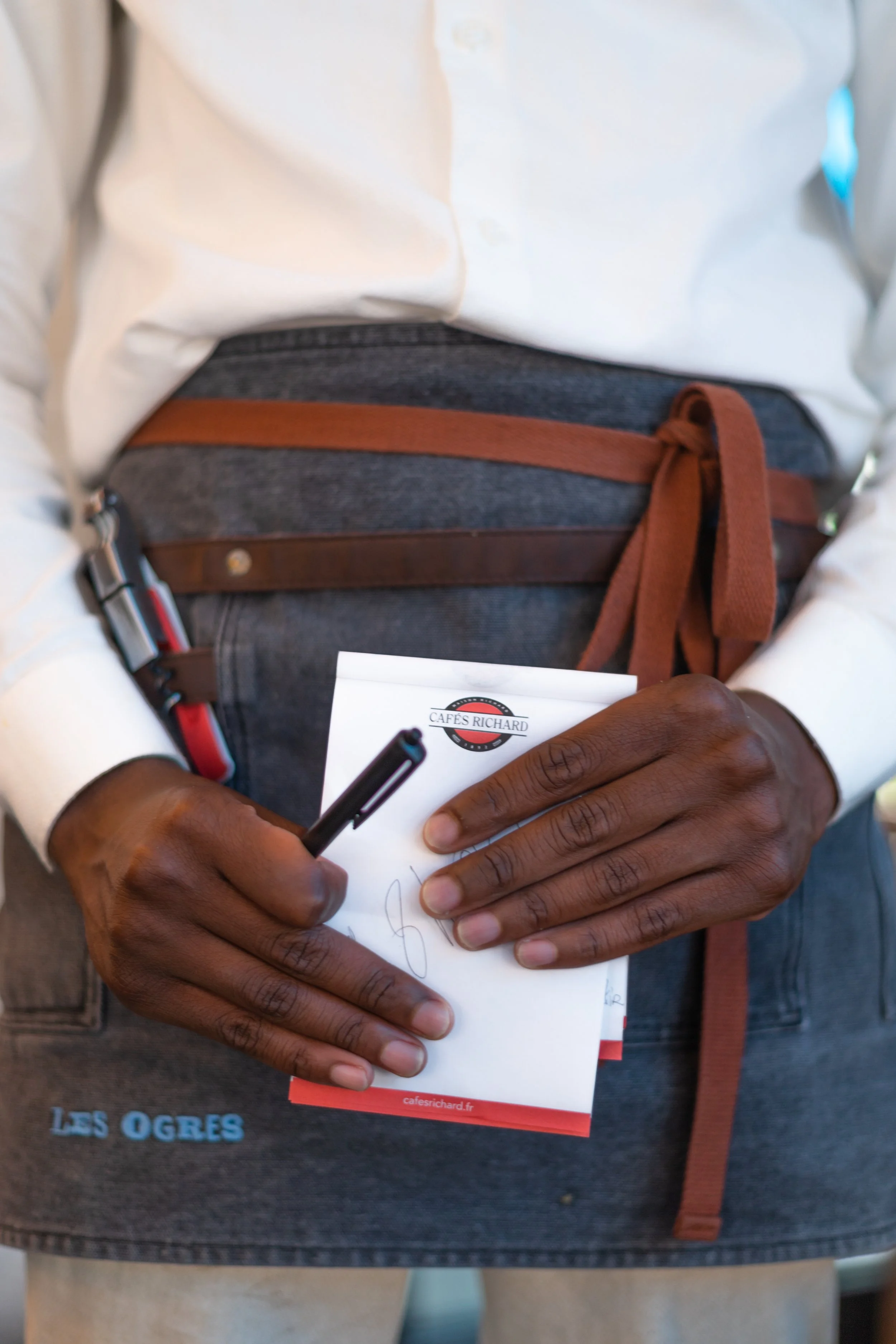 A person wearing a white shirt and an apron with a logo from Cafe Richard, holding a small notepad and a black pen, standing with hands close together.