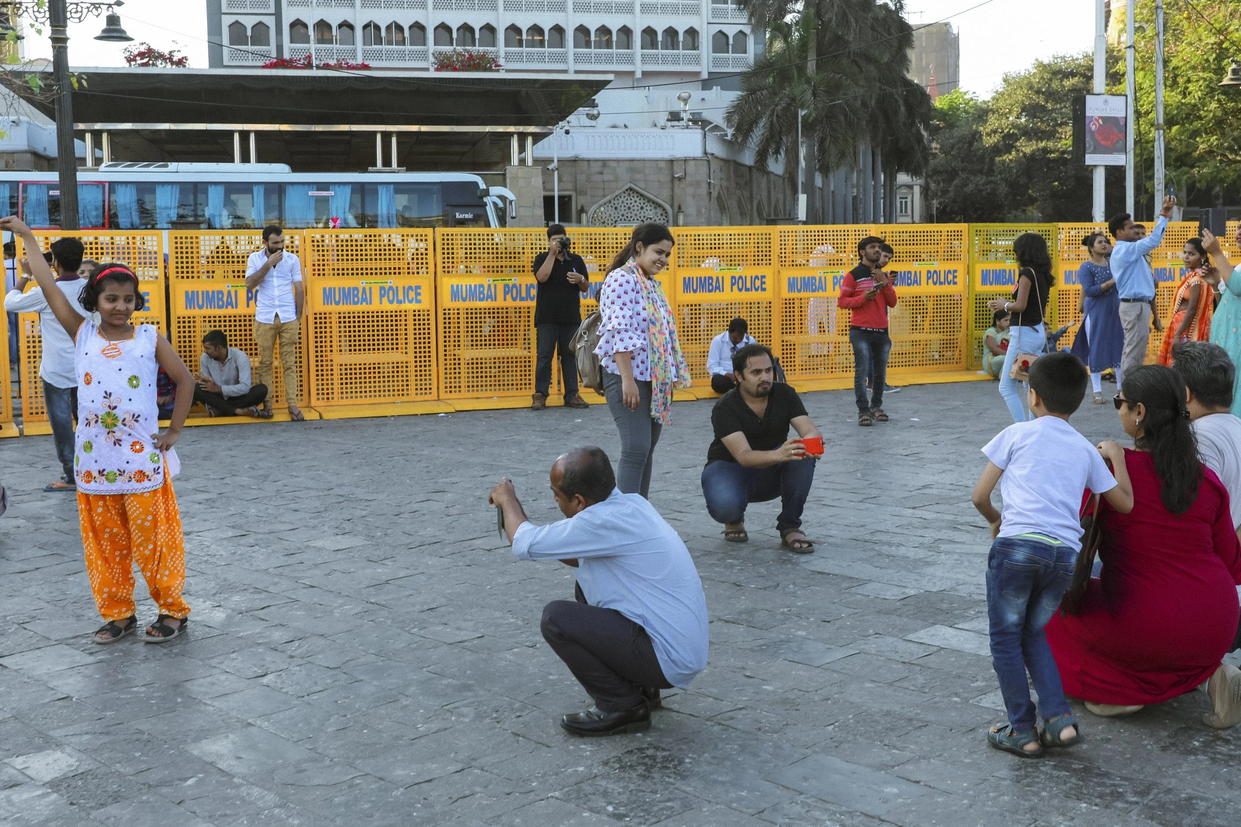 People taking photographs and playing in a public square in front of a yellow barricade labeled 'Mumbai Police,' with trees and buildings in the background.