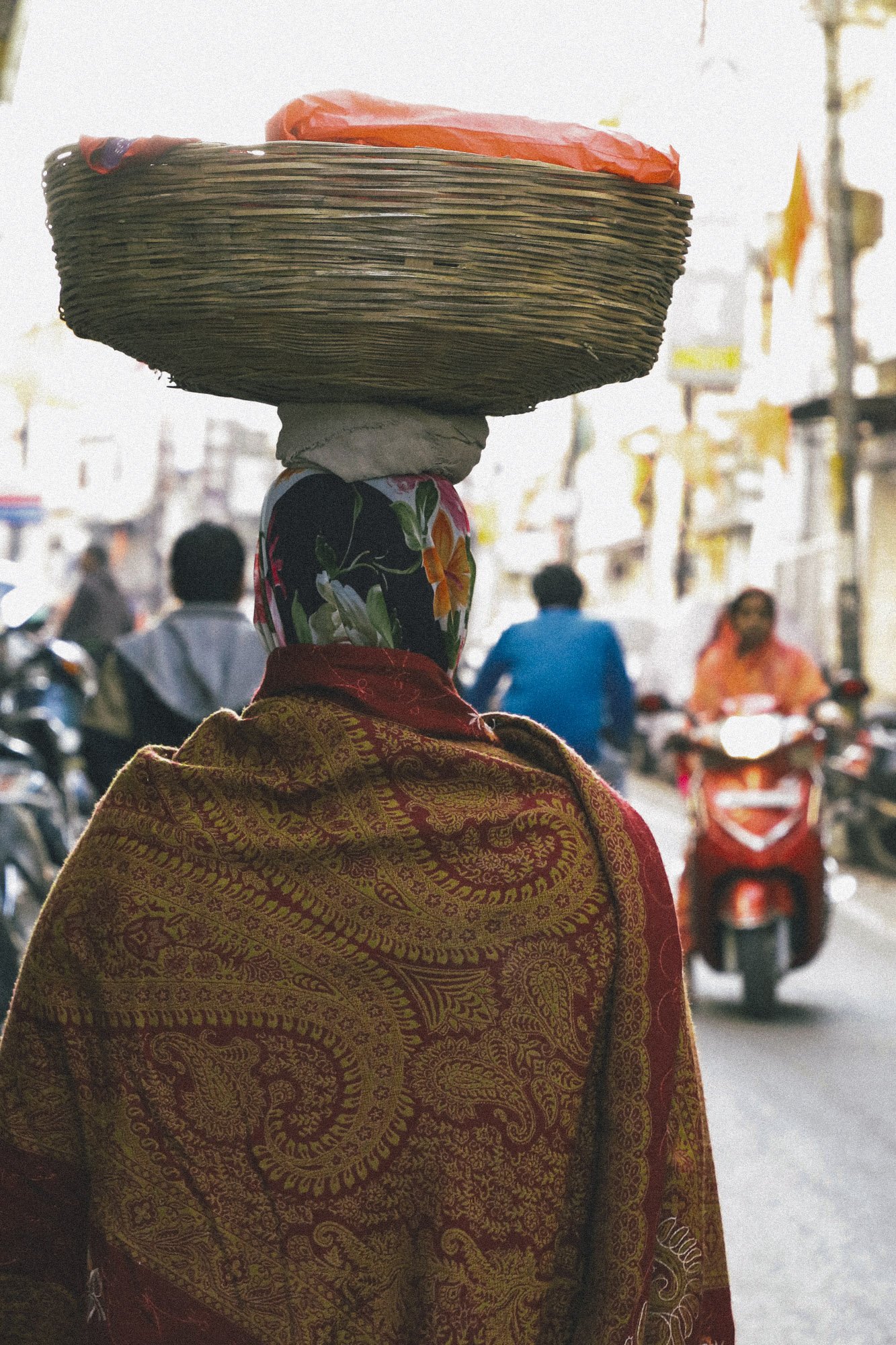A woman walking on a busy street carrying a large basket on her head, with several motorcycles and other people in the background.