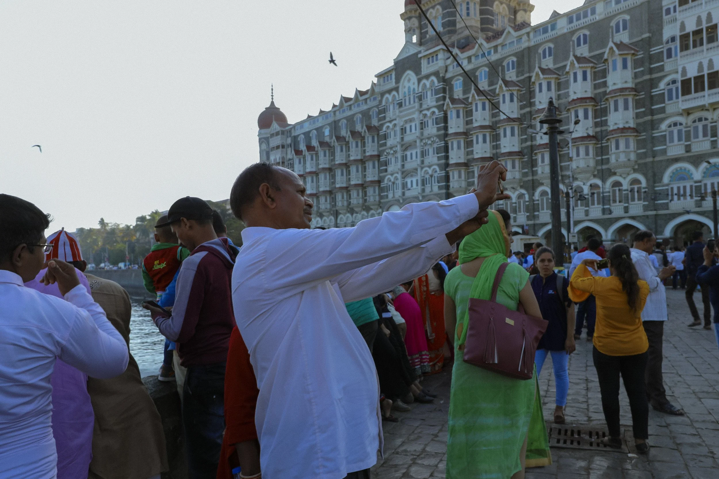People gathered outdoors near a riverfront with a large, ornate building in the background. One man is taking a photo with his phone, and others are standing and talking. Some are wearing traditional Indian attire and colorful clothing. Birds are fly