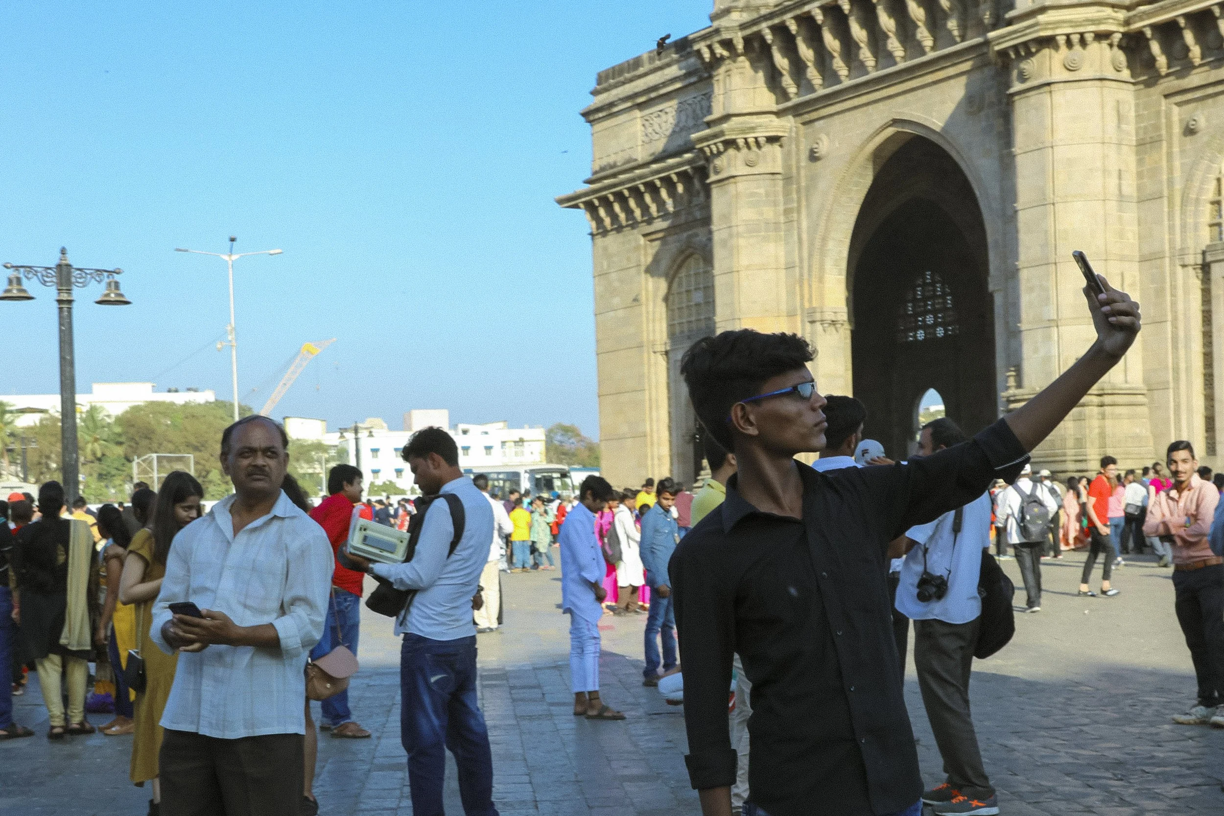 Young man taking a selfie in front of the India Gate, with other people walking and standing nearby at the monument site on a sunny day.
