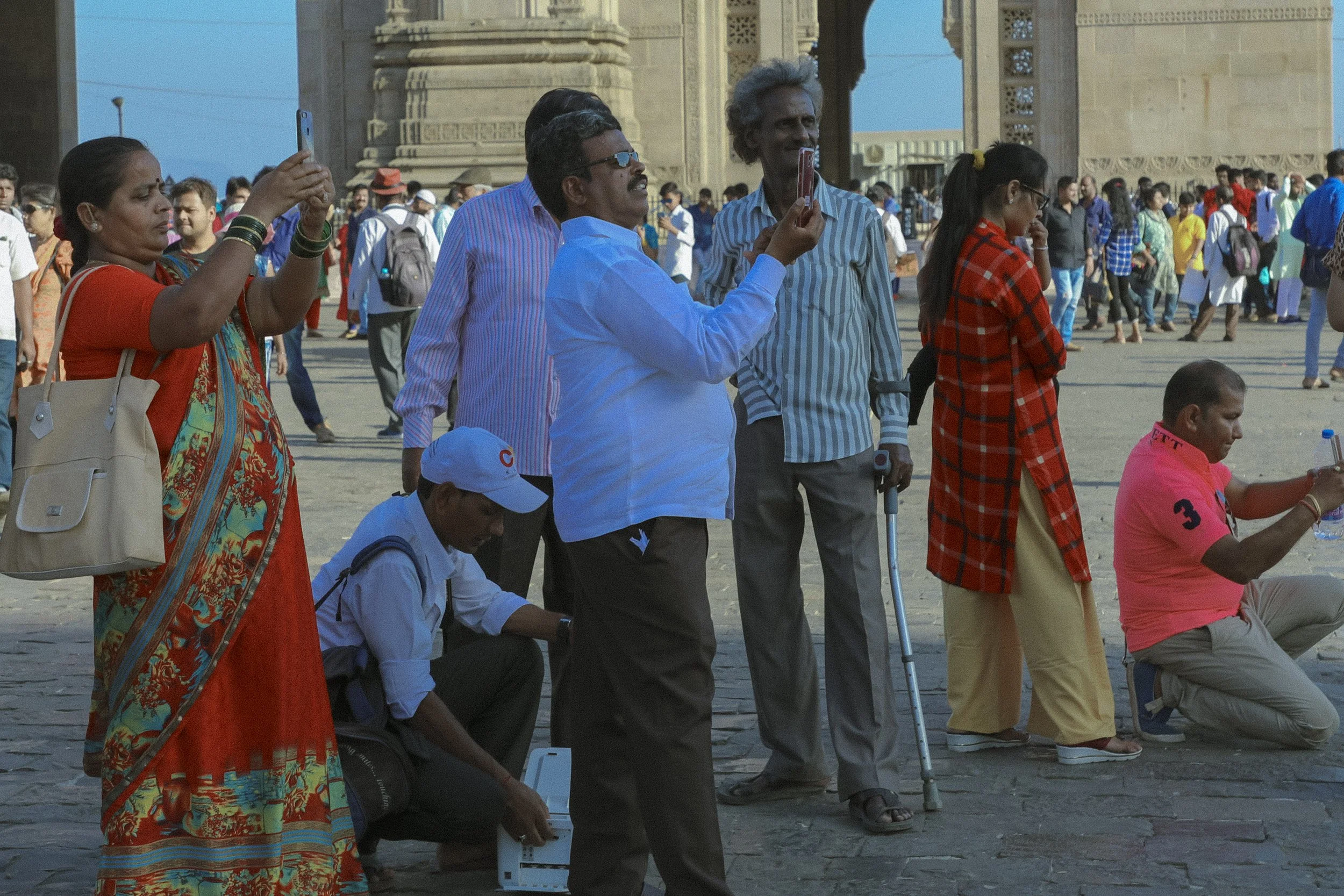 A group of people in front of a historical monument, some taking photos, others sitting or standing, with a crowd in the background.
