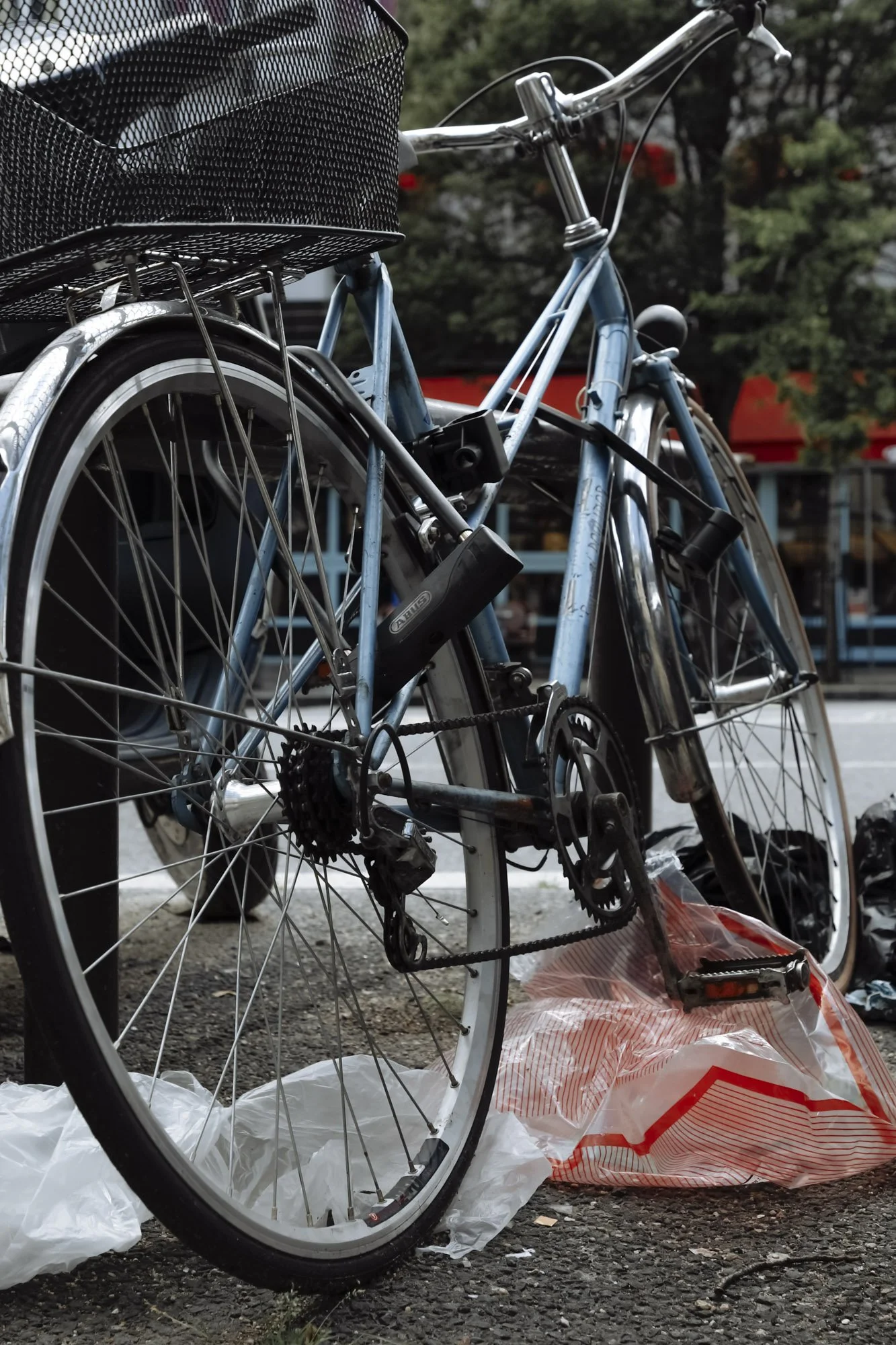 A blue bicycle with a black basket on the front, parked on a street. The bike appears to be damaged or discarded, with some bags nearby.