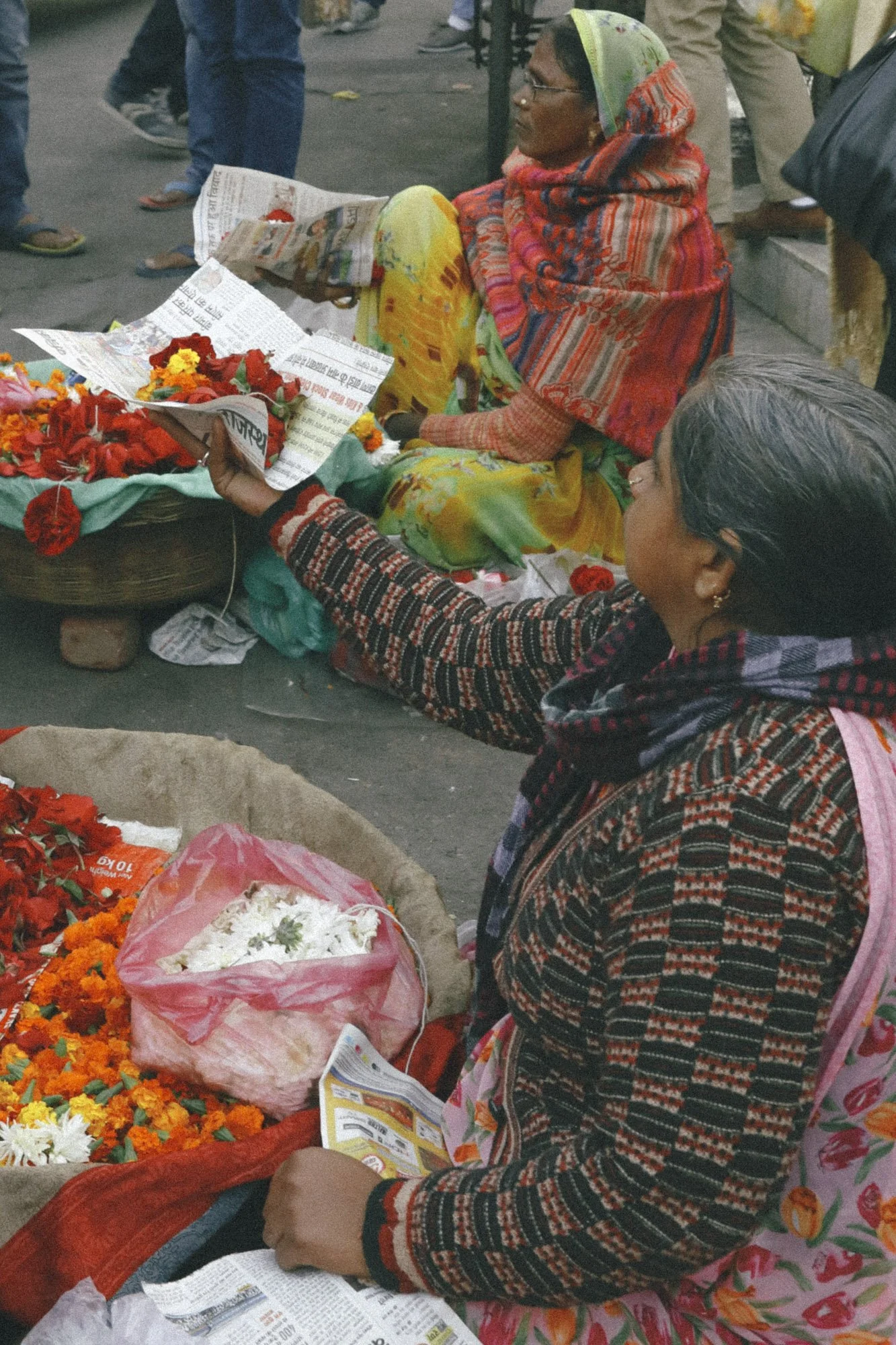 Two women sit on the ground selling flowers and reading newspapers, with people walking in the background.