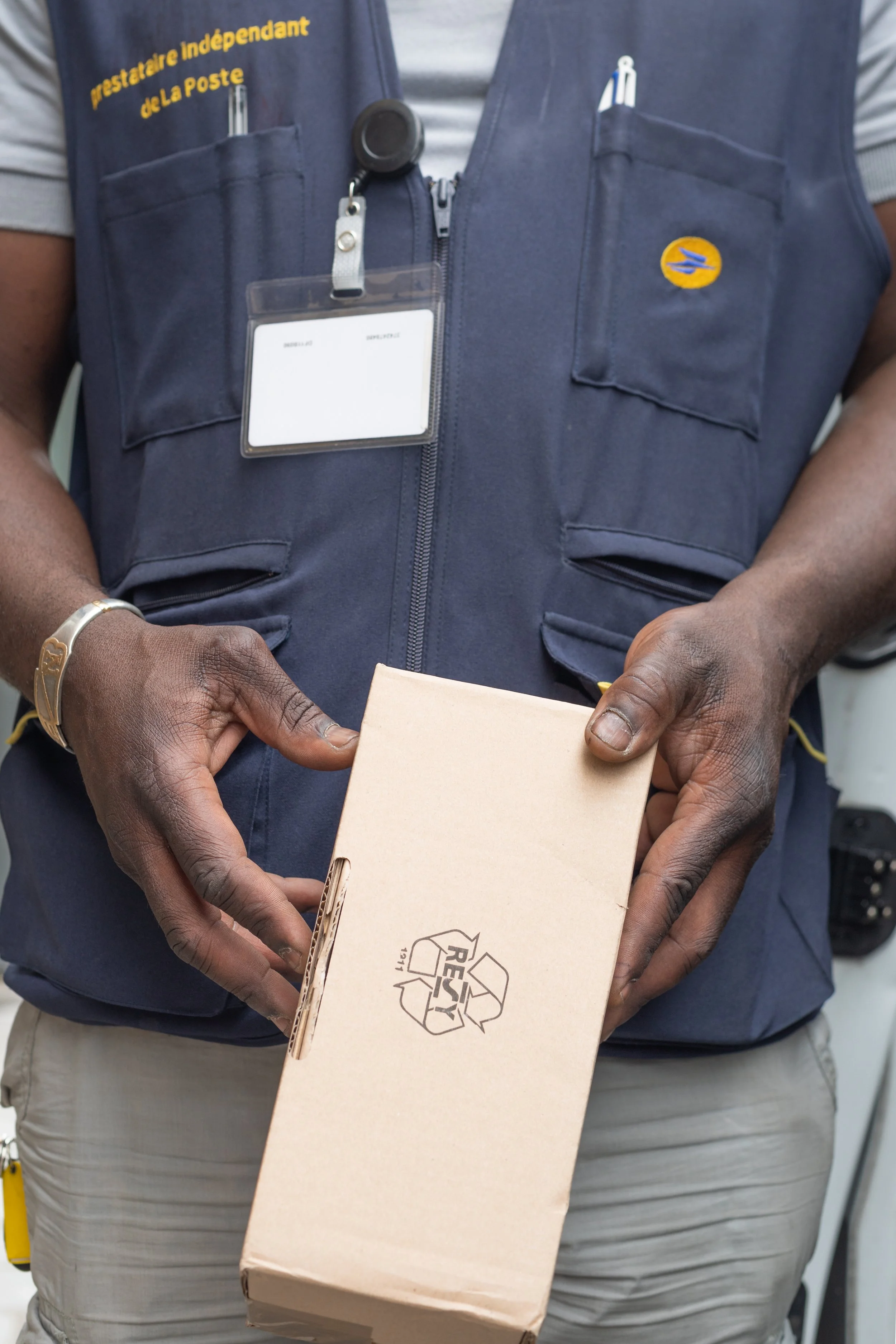 A postal worker holding a small cardboard box with a recycling symbol on it, wearing a blue vest with postal insignia and a name badge, with a pen in the vest pocket.