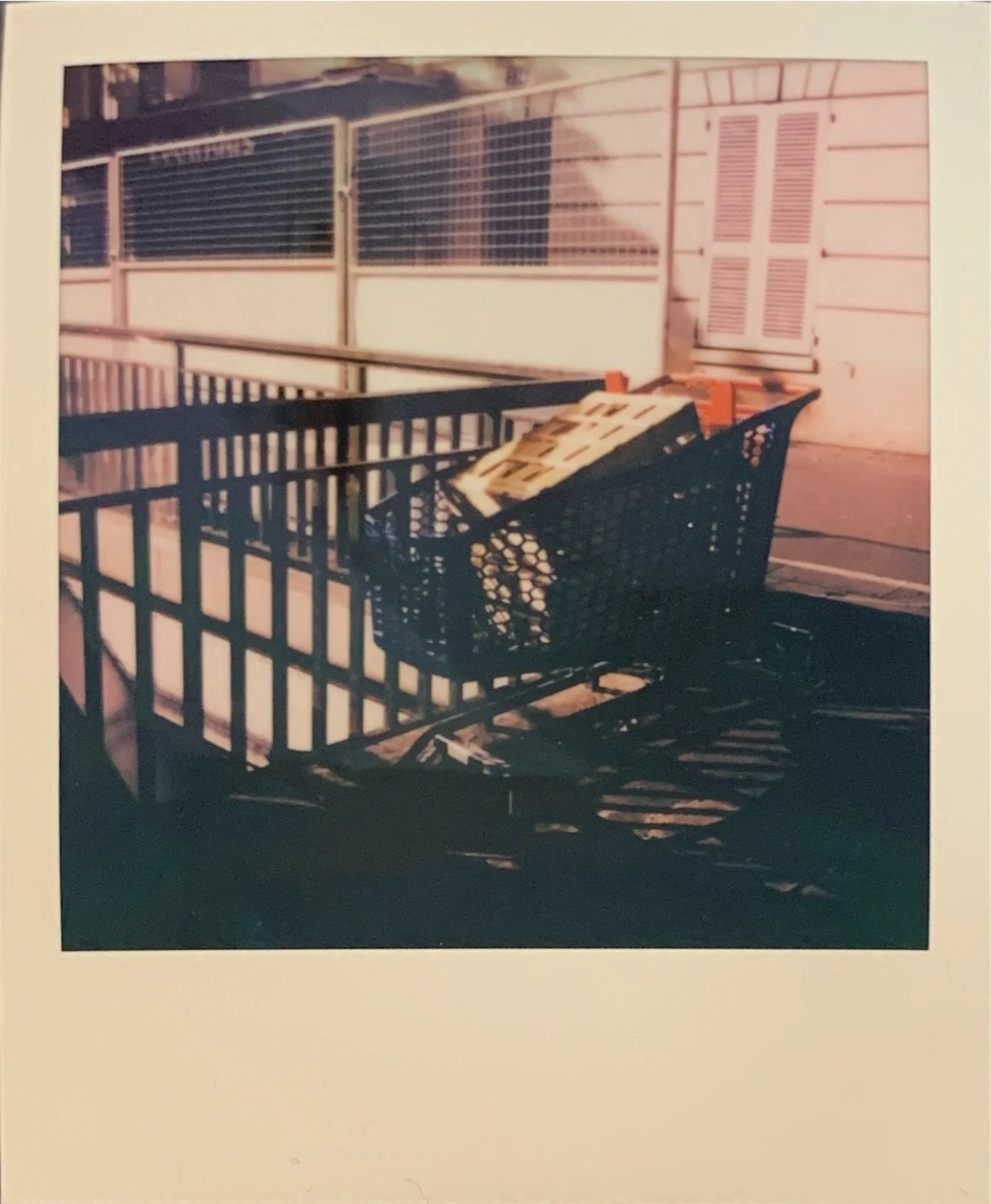 A shopping cart filled with boxes is on a balcony or outdoor area with metal railings, with a building and closed shutters in the background.