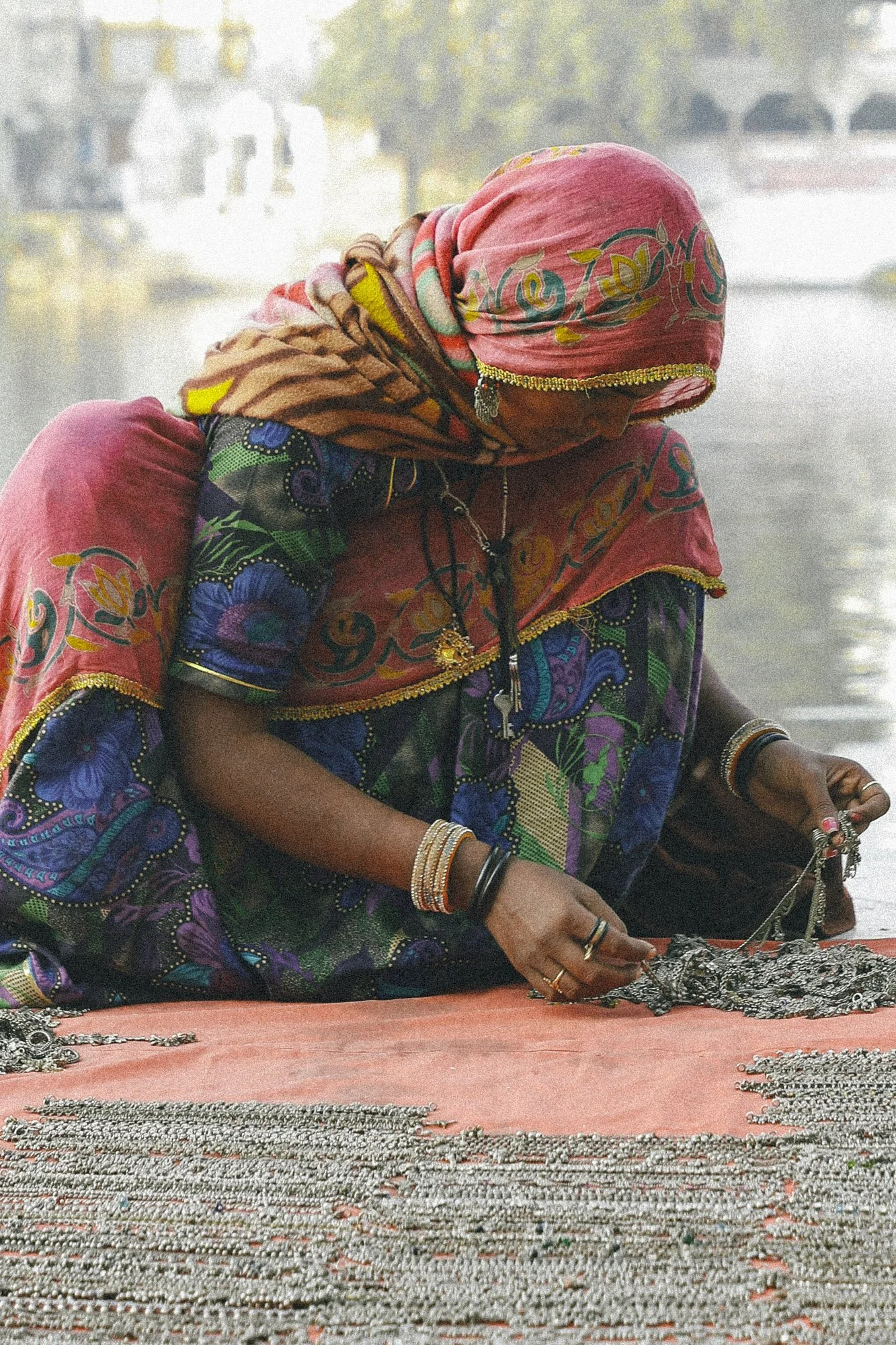 A woman dressed in colorful traditional attire with a headscarf, sitting at a market stall and arranging jewelry such as necklaces and bracelets.