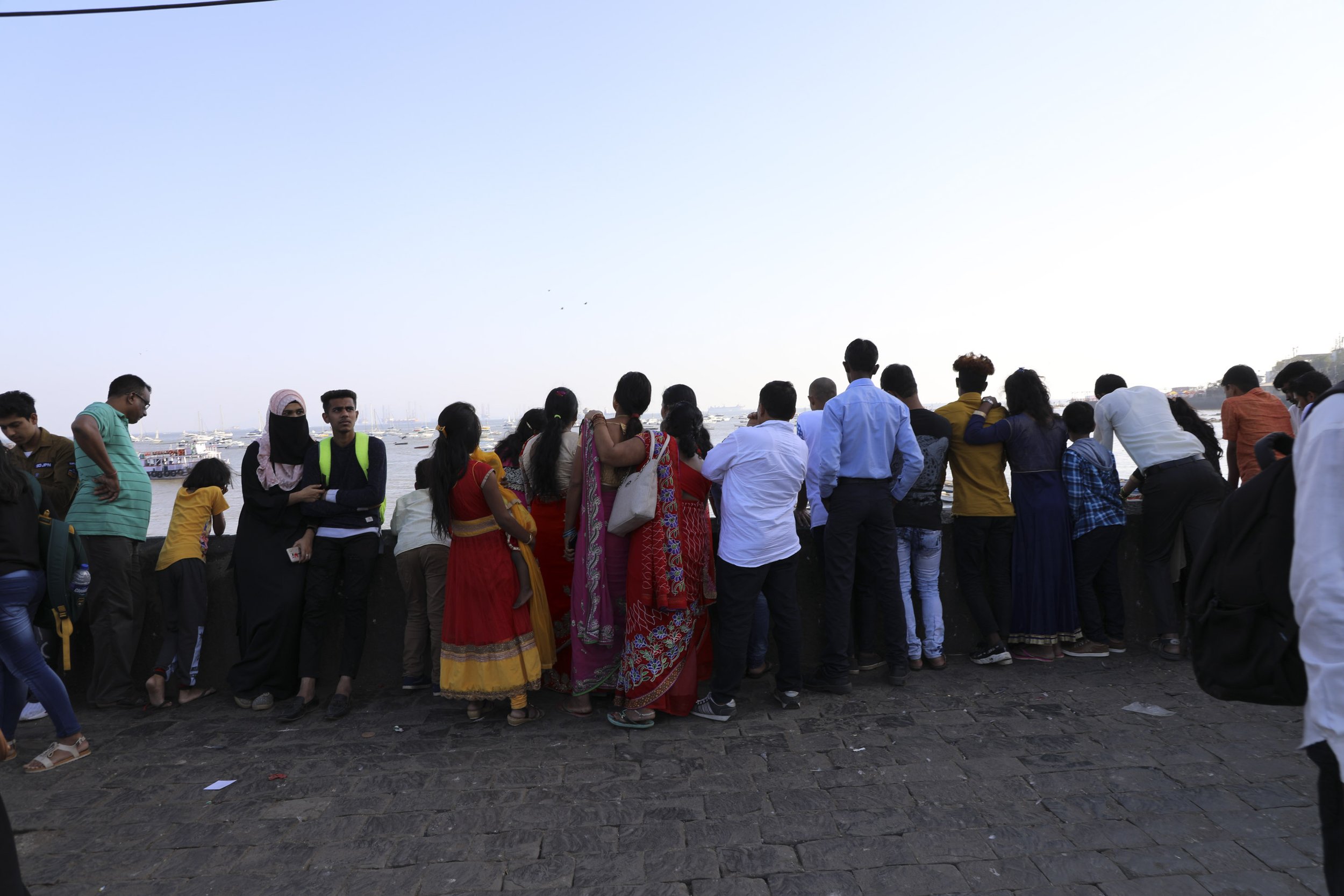 Group of people standing along a waterfront, looking out at the boats on the water, during daytime with clear sky.