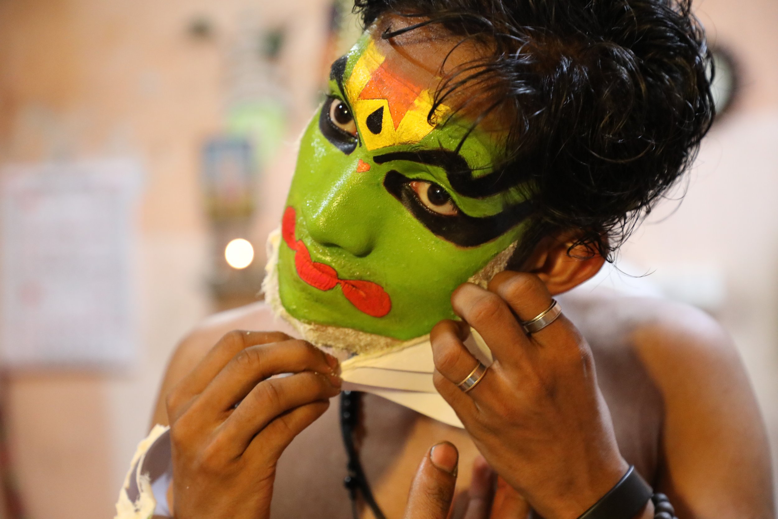 Person with vibrant green face paint, black and red detailing, wearing a mask or headpiece, adjusting it with their hands.
