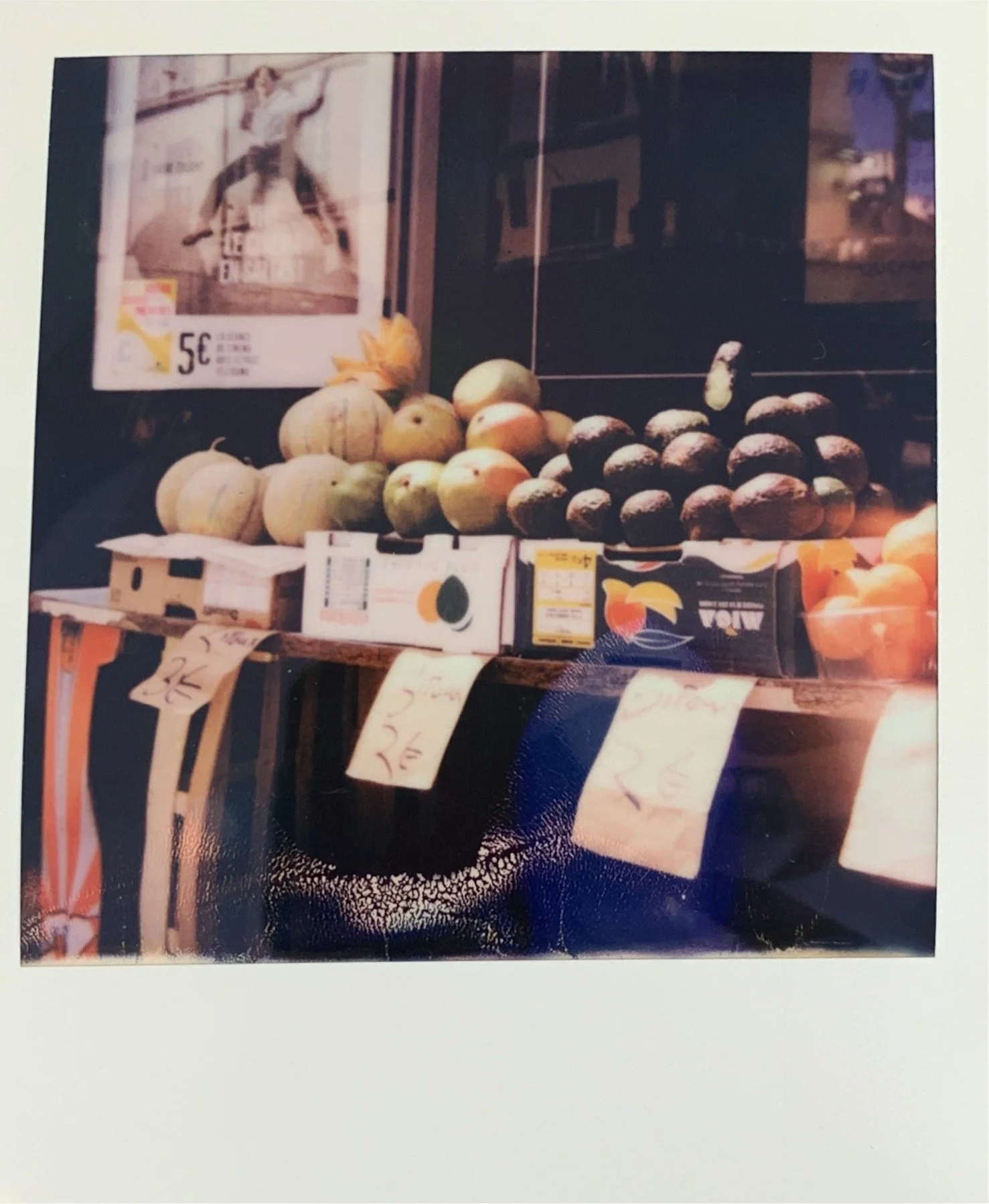 Market stand with various melons and fruit on display, including cantaloupe, honeydew, and some darker melons in boxes, with handwritten price signs hanging in front.