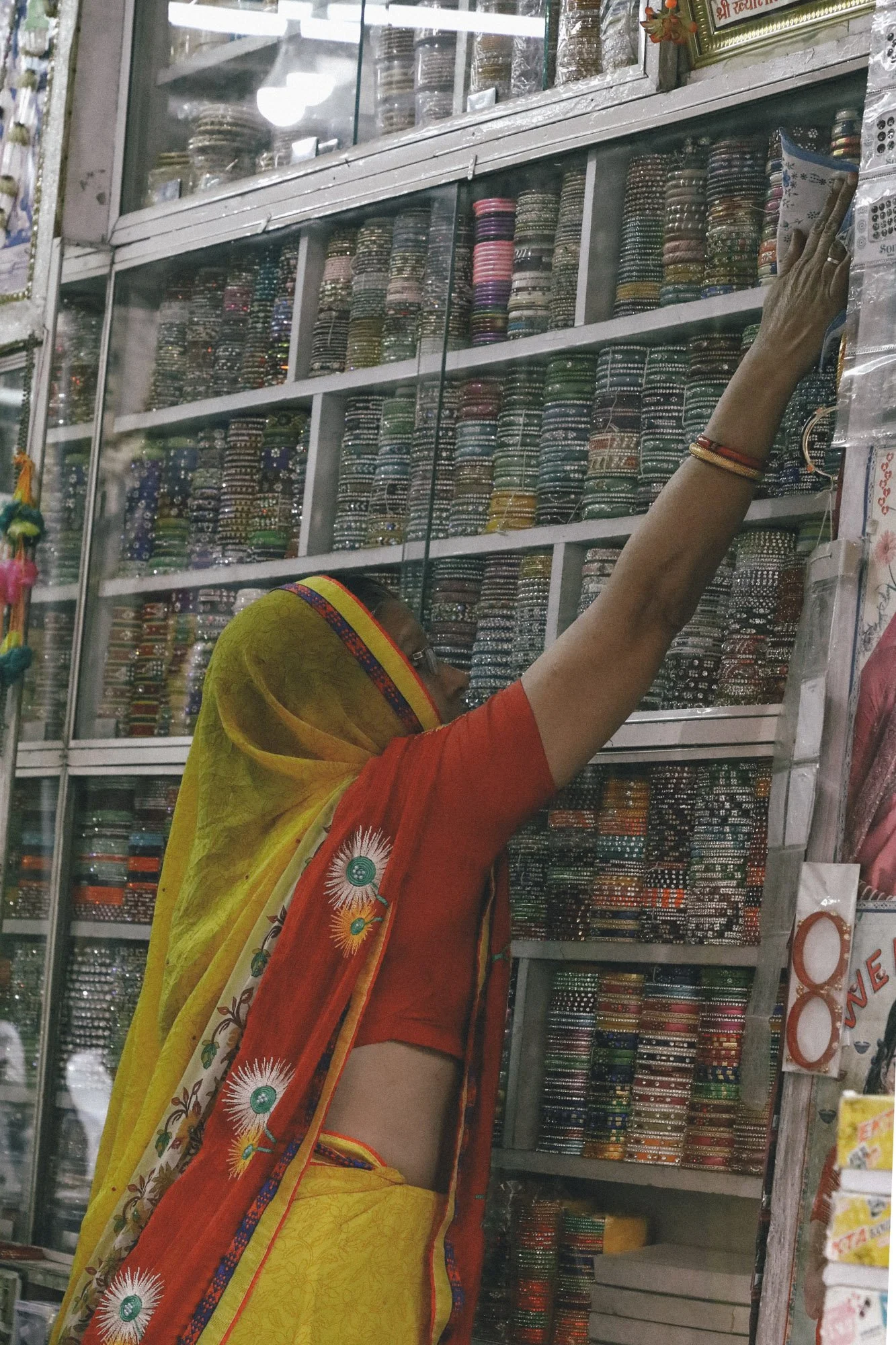 A woman wearing a yellow saree with a red blouse and a red scarf shopping for bangles at a jewelry shop with shelves full of colorful bangles.