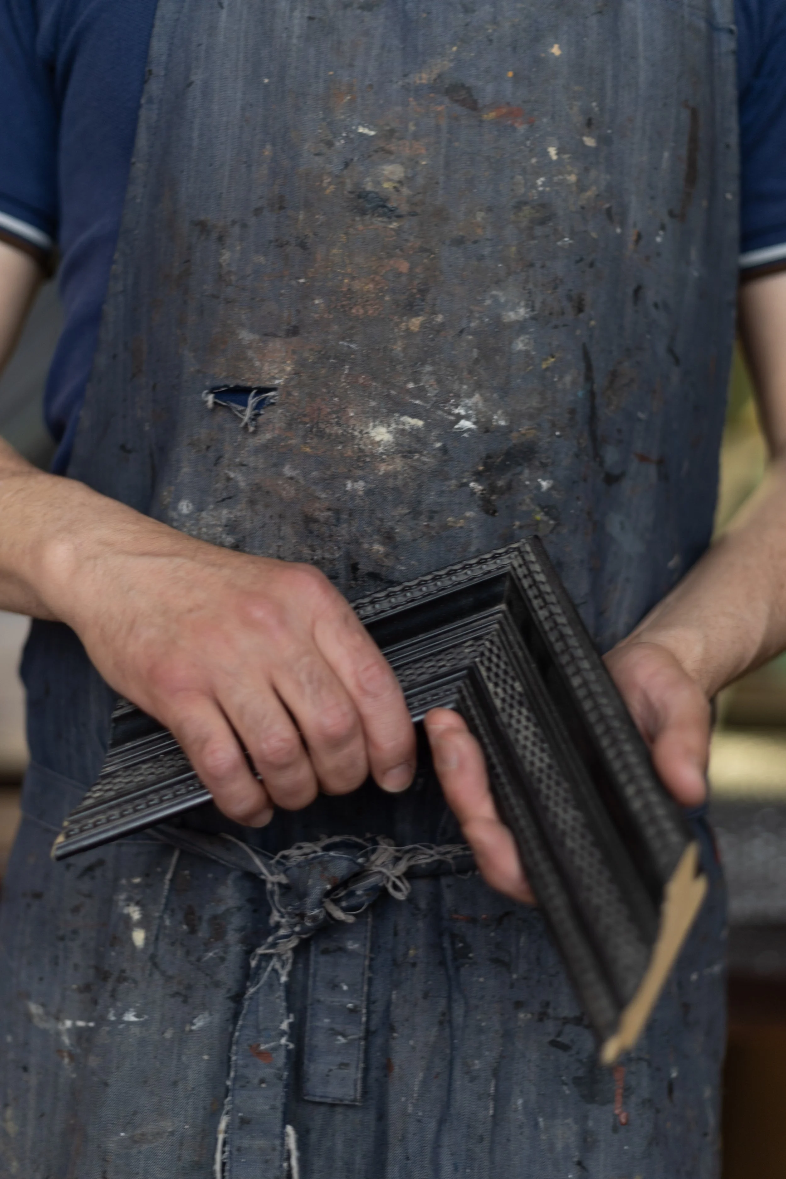 A craftsman in a stained, paint-splattered apron holding a black, textured picture frame.