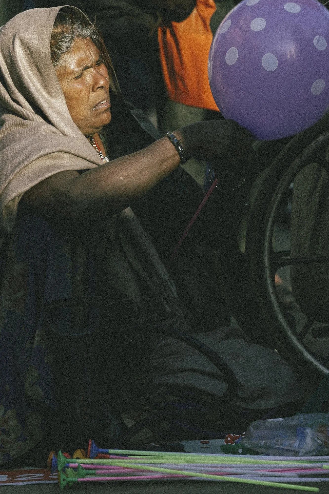 An elderly woman in a beige hoodie and black gloves is sitting in a car, reaching out to receive a purple balloon with polka dots from someone outside the car. The scene appears to be at a nighttime gathering with various colorful objects and people 