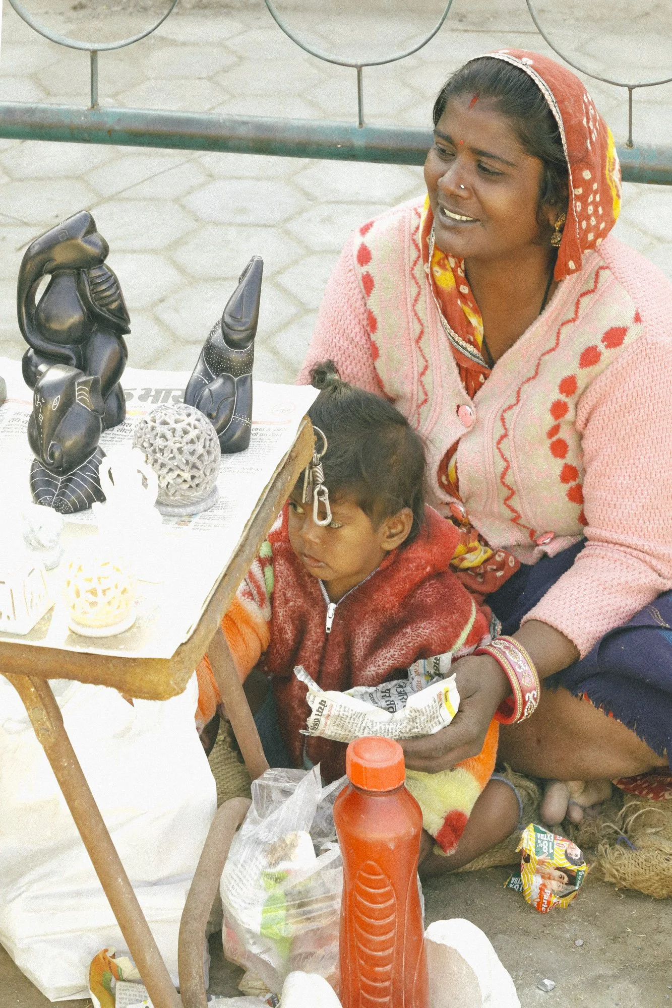 A woman with traditional Indian attire and jewelry sitting next to a young girl with a macrame bracelet on her forehead, at a street stall with carved black figurines, a spherical decorative object, and snacks.