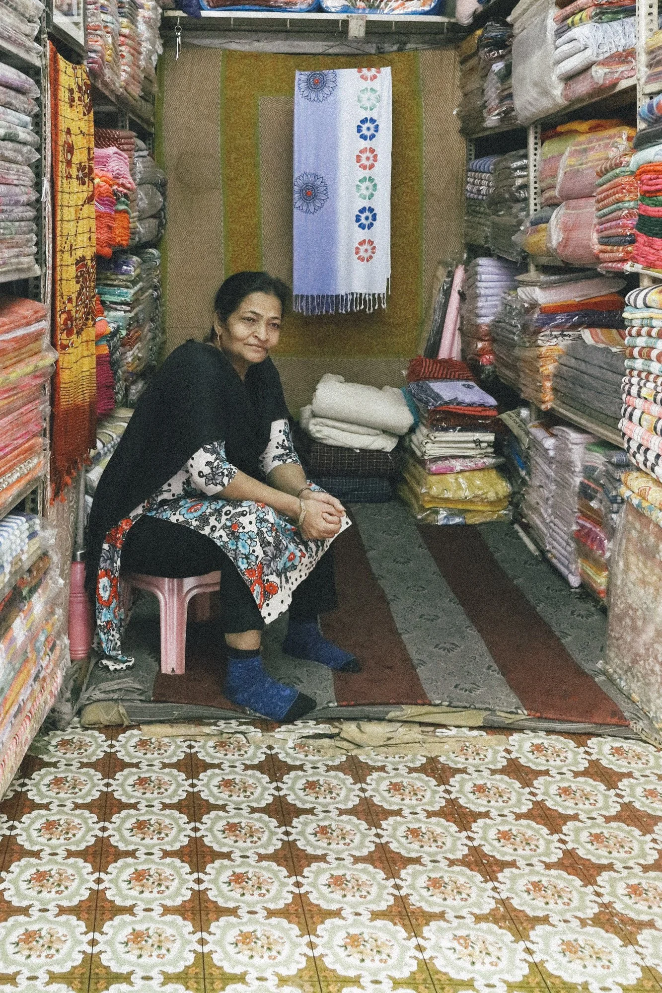 A woman sitting on a pink stool inside a fabric shop, surrounded by shelves filled with colorful fabrics and textiles.