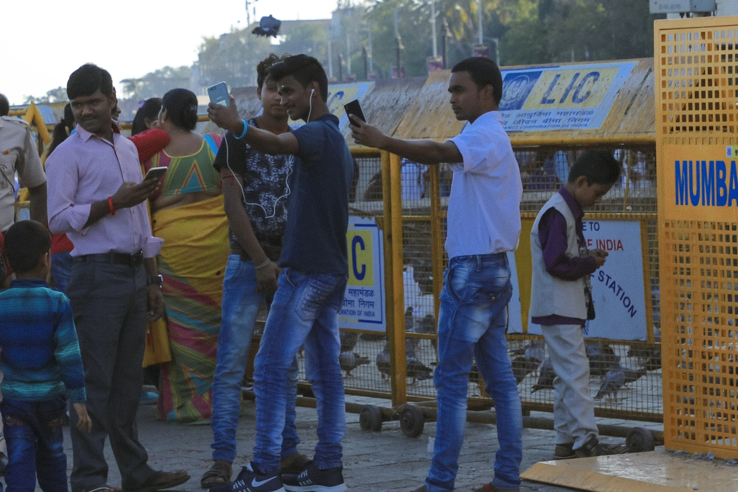 Group of people, including young men and children, gathered near a metal barrier at a transport station in India. Some are using smartphones, with one wearing earphones, and a person is dressed in traditional Indian attire. The background shows signs