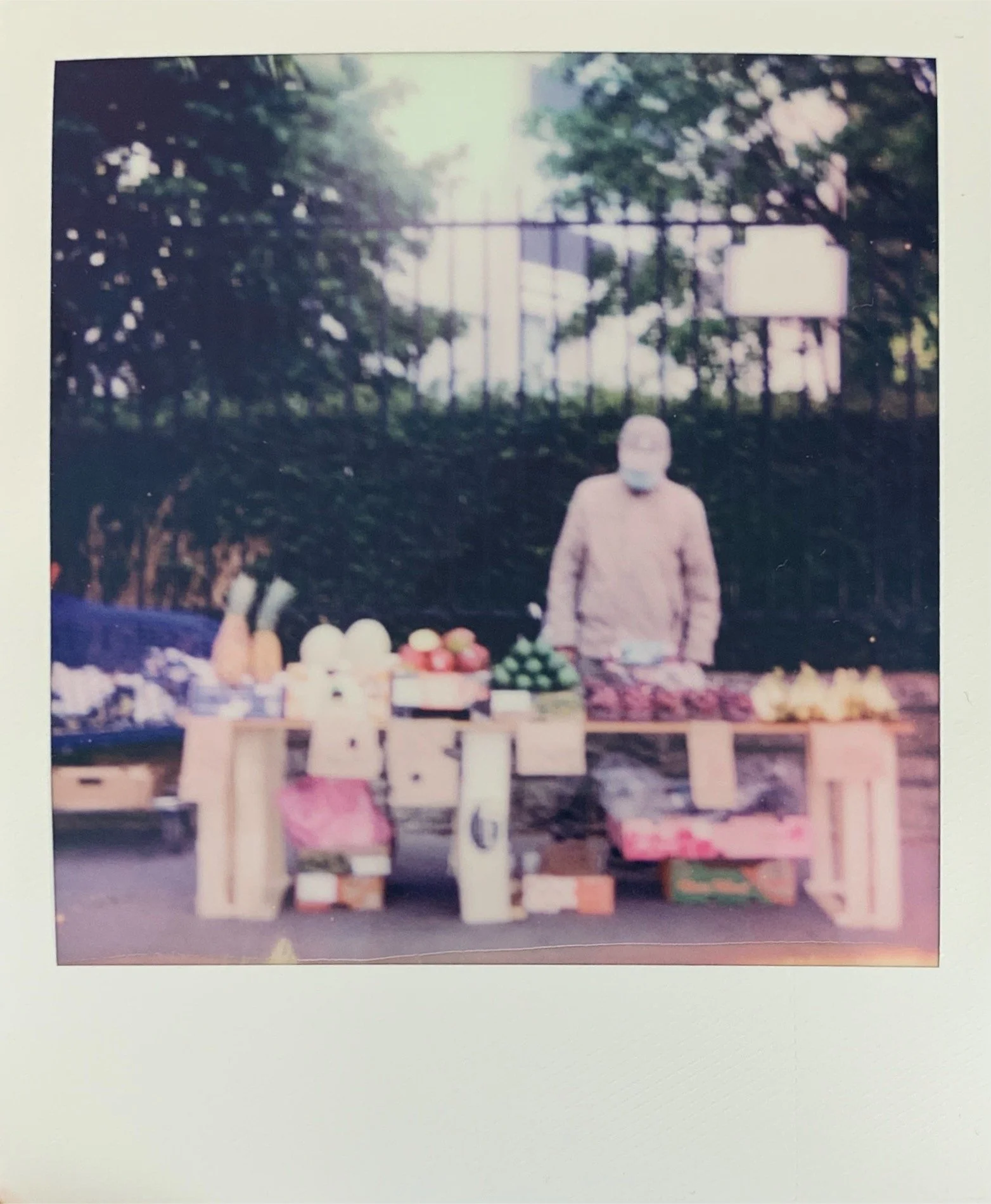 A person wearing a face mask standing behind a fruit and vegetable stall outdoors, with trees and a fence in the background.