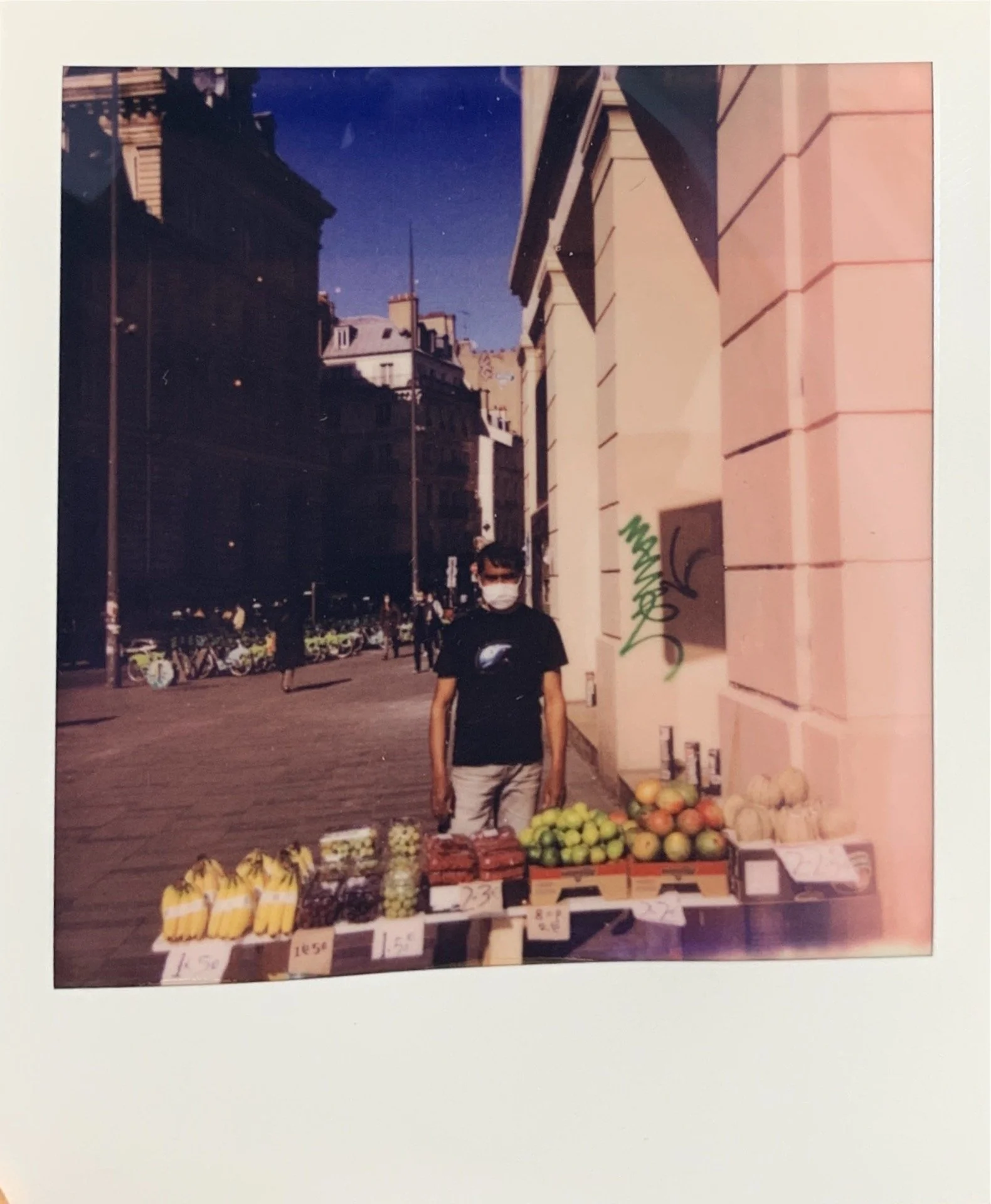 A person wearing a mask standing behind a street fruit stand with bananas, grapes, apples, and melons on display, in an urban setting with buildings and bicycles in the background.