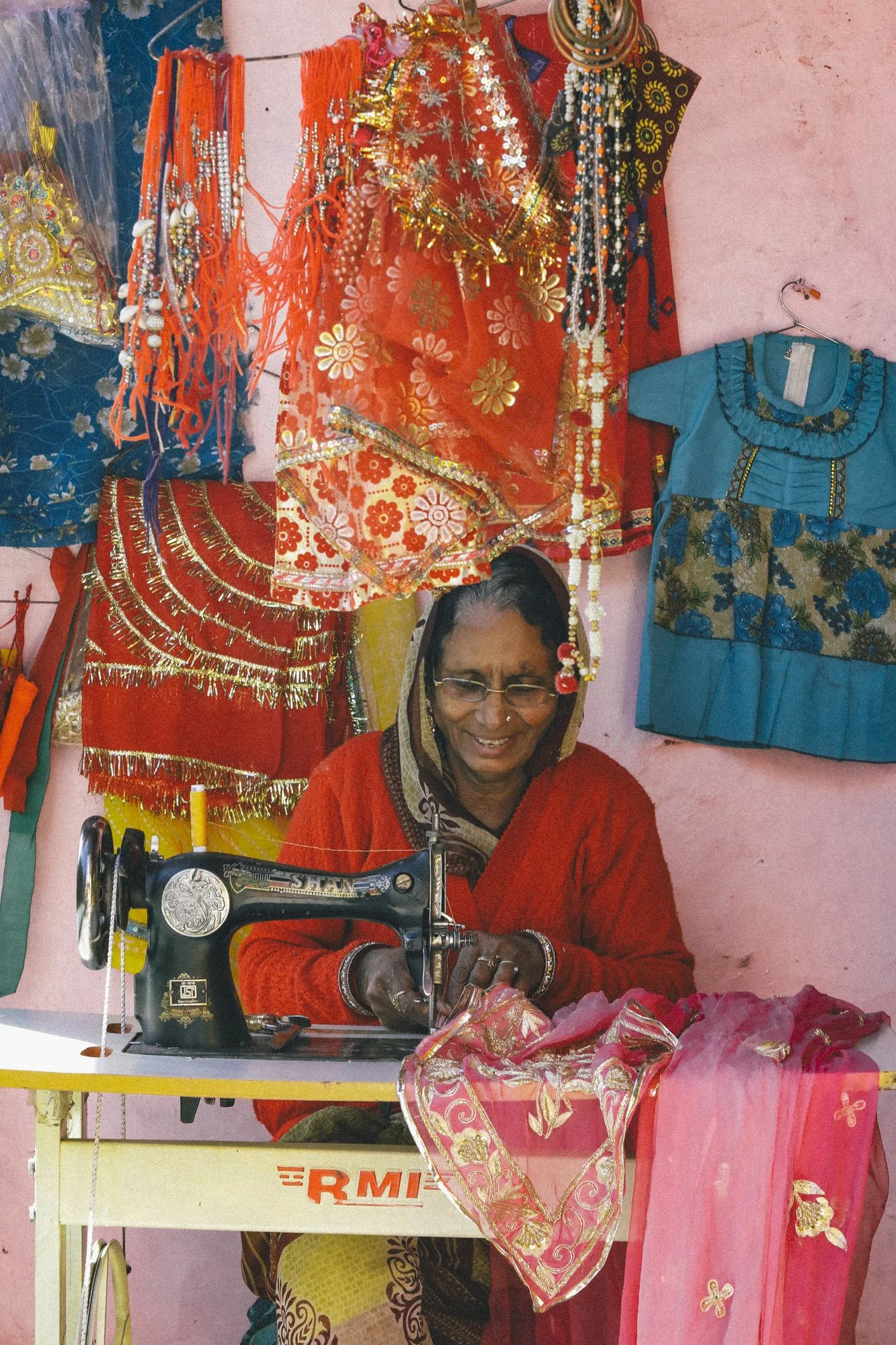 An elderly woman sewing pink and gold embroidered fabric on a vintage black Singer sewing machine, with colorful traditional garments hanging on a pink wall behind her.