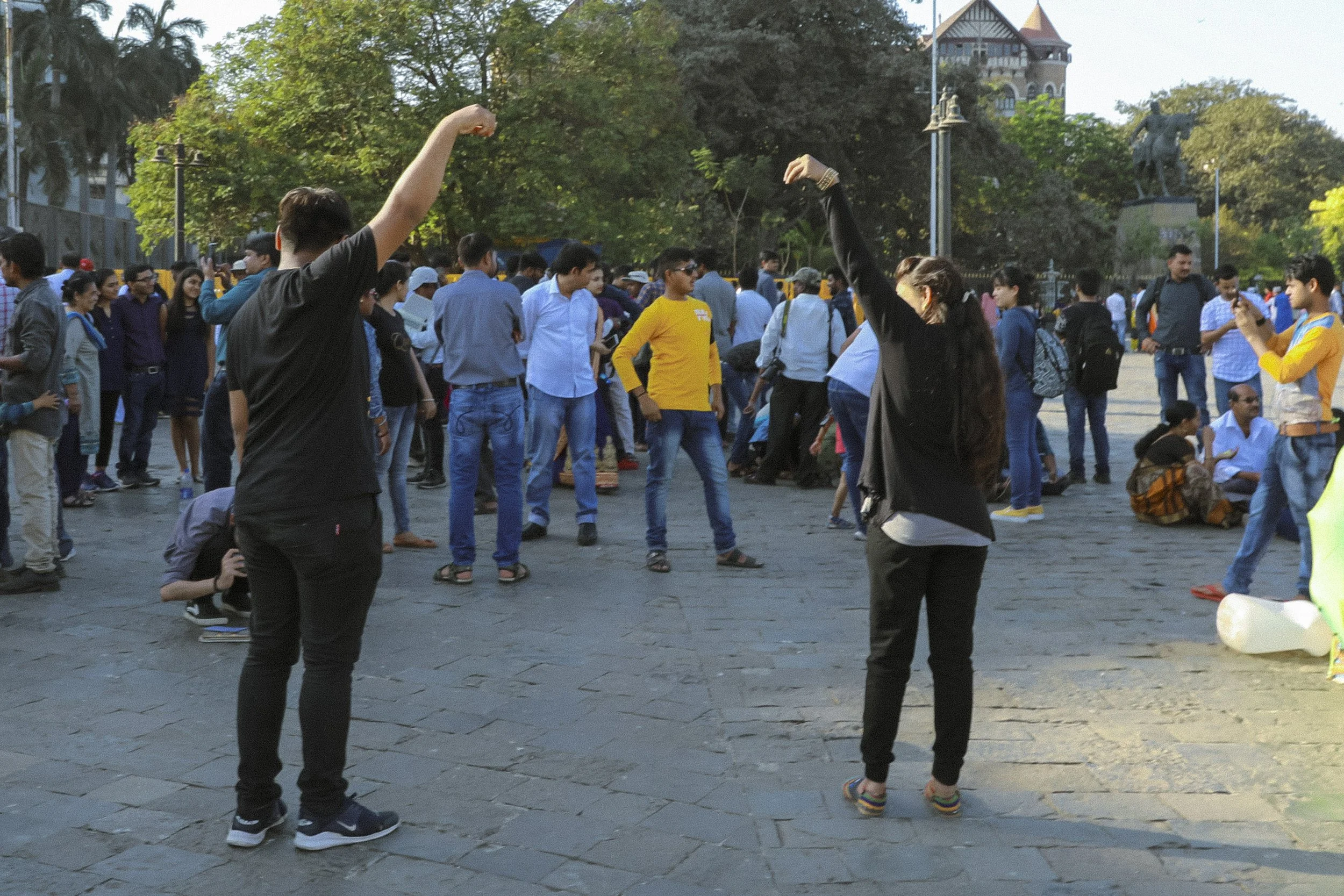 A group of diverse people gathered outdoors in a park or plaza with trees and historic buildings in the background. Some people are dancing, taking photos, or sitting on the ground, enjoying a social event.
