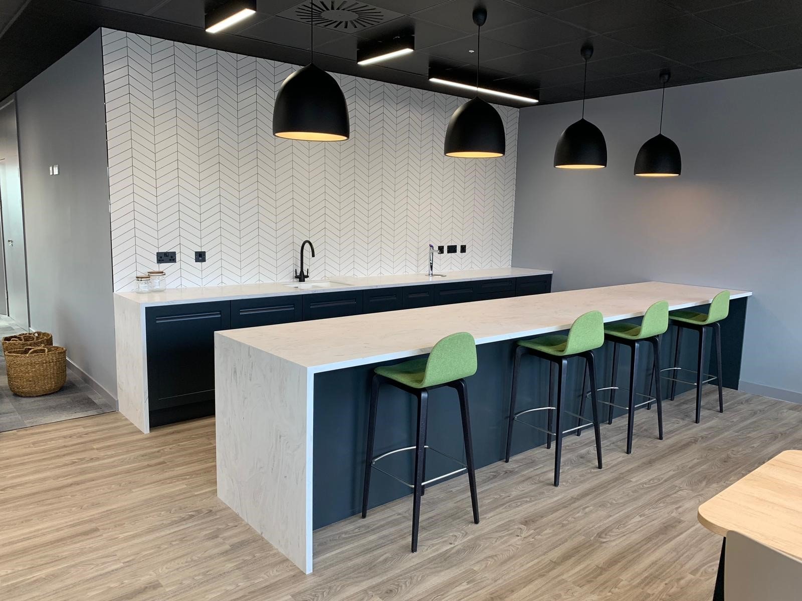 Modern kitchen with a long counter, green bar stools, pendant lights, and a herringbone tiled backsplash, part of an office fit-out by GM Projects.