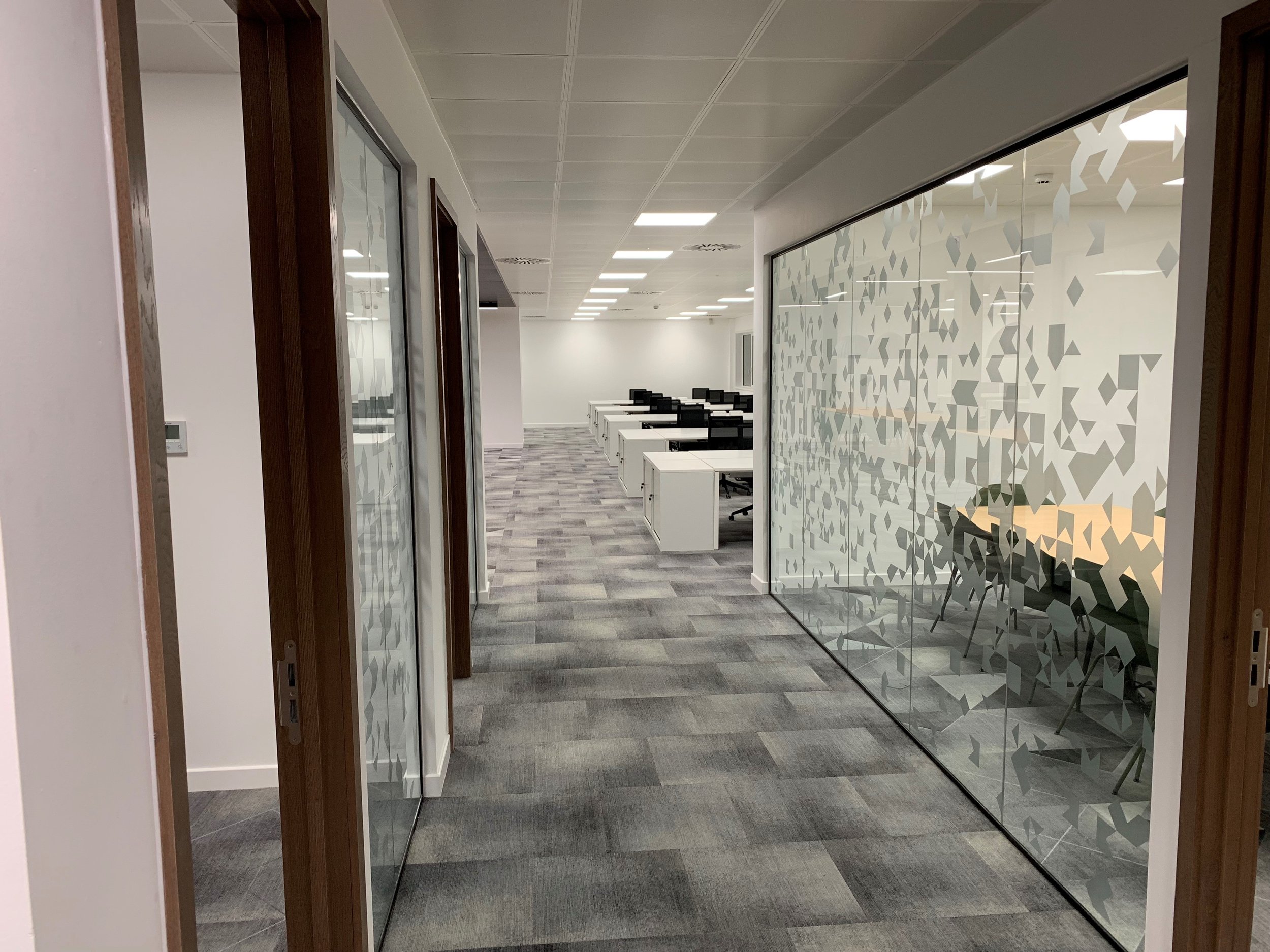 Modern office hallway with glass partitions and geometric patterns, carpeted flooring, and cubicles in the background, part of an office fit-out by GM Projects.