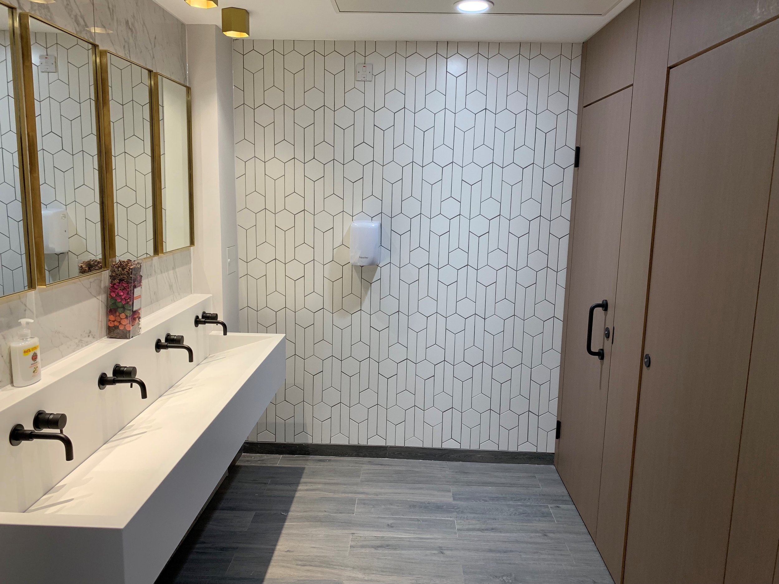 Modern bathroom with geometric tile wall, three sinks with black faucets, and large mirrors, part of an office fit-out by GM Projects.