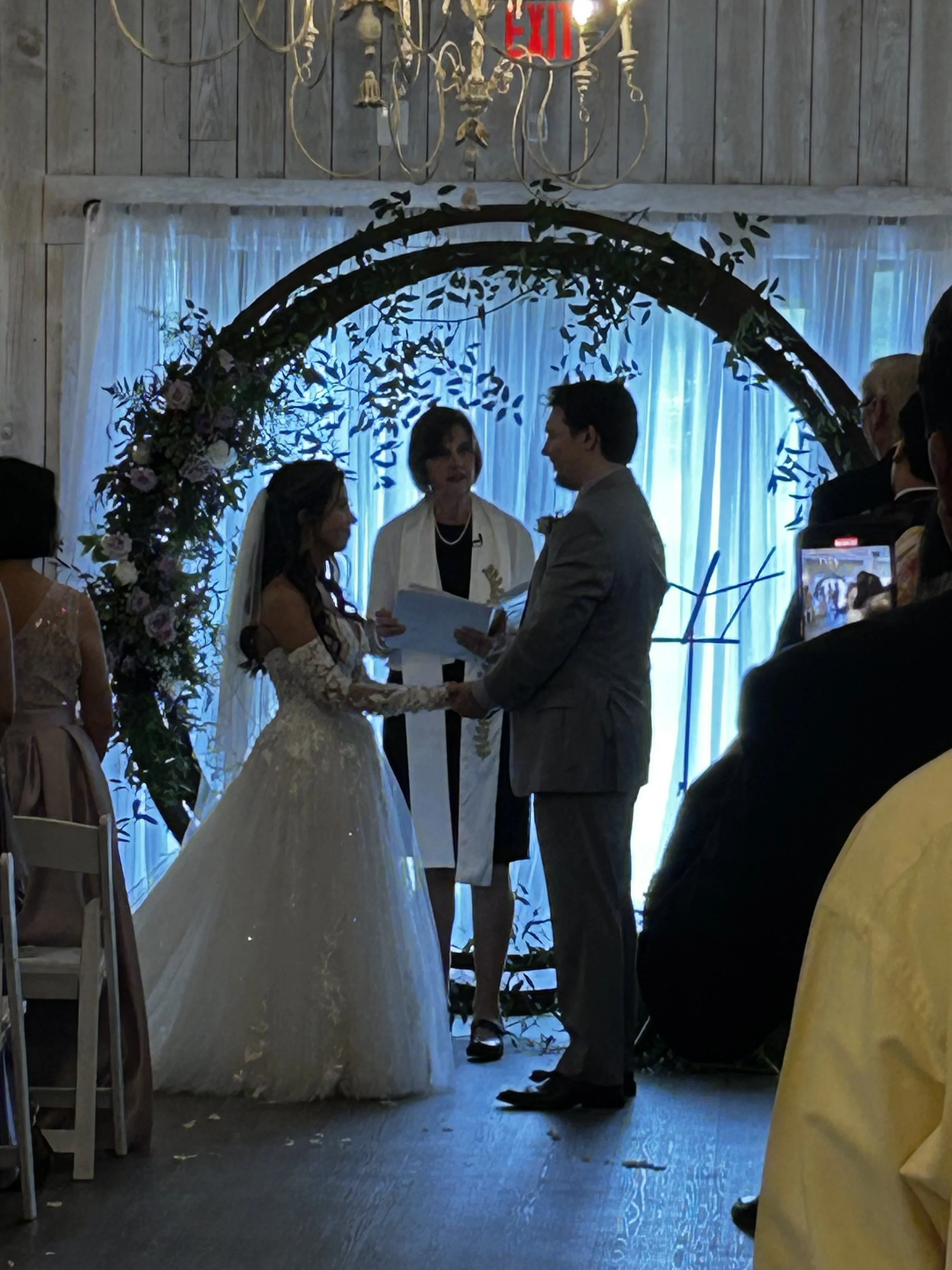 A wedding ceremony with a bride and groom holding hands, standing under a decorated arch, facing each other with an officiant behind them. Guests seated on either side, with a blue-lit curtain in the background.
