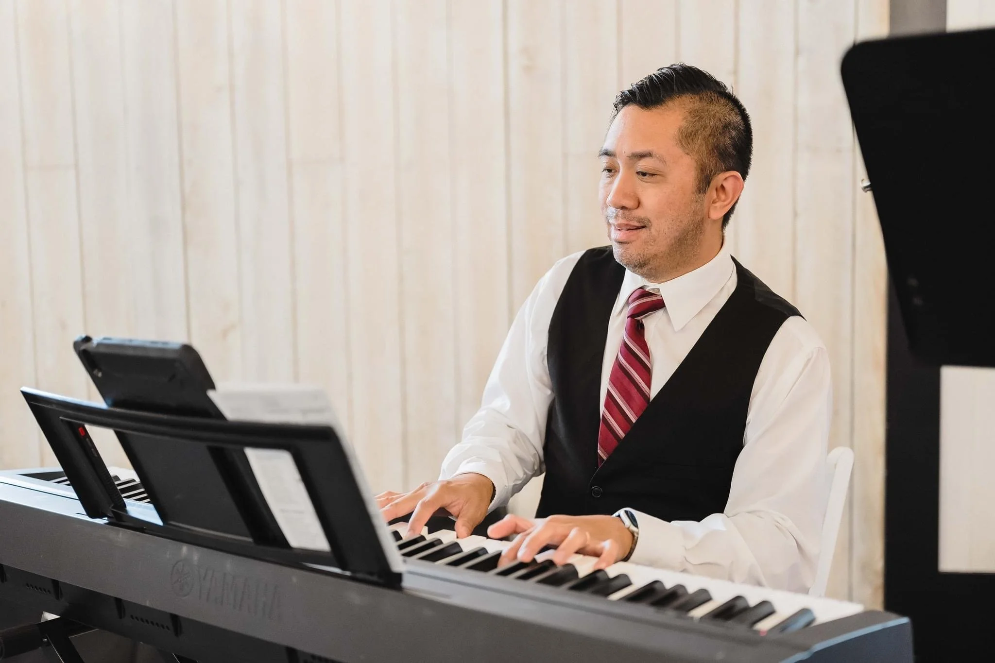 A man in a white shirt, black vest, and red striped tie playing a Yamaha digital piano in front of a wooden wall.