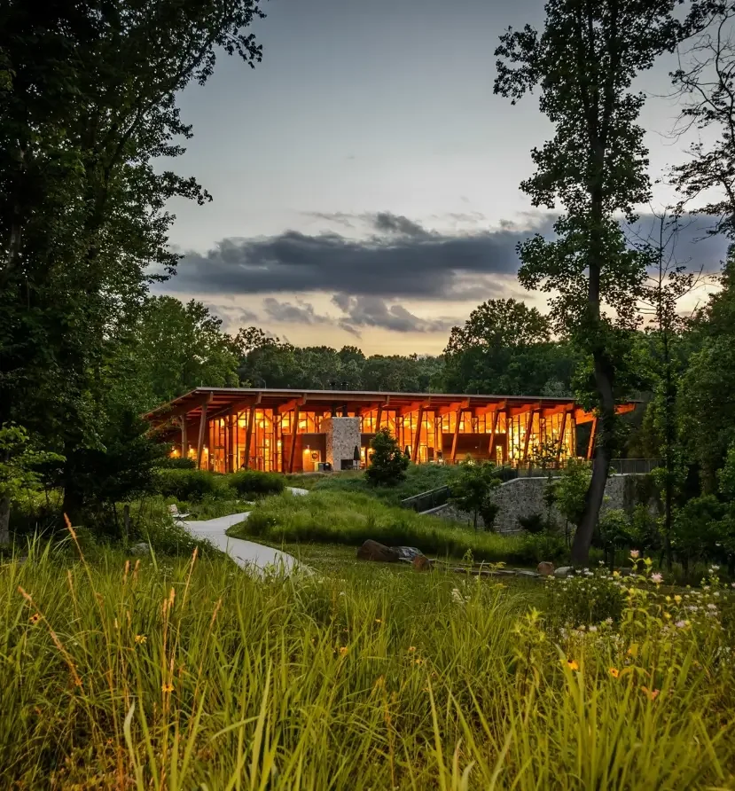A modern building with warm interior lighting, surrounded by green trees and tall grass, under a cloudy sky at dusk.