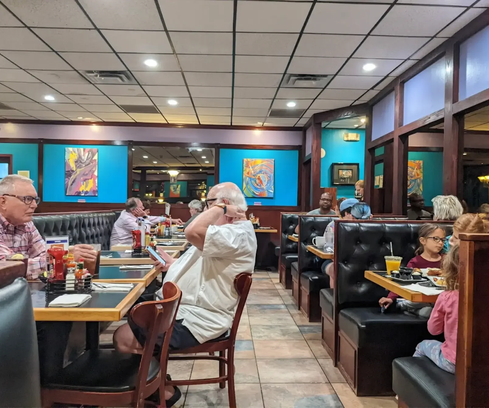 A busy restaurant with customers seated at tables and booths, colorful abstract paintings on the blue walls, and a tiled ceiling with recessed lighting.
