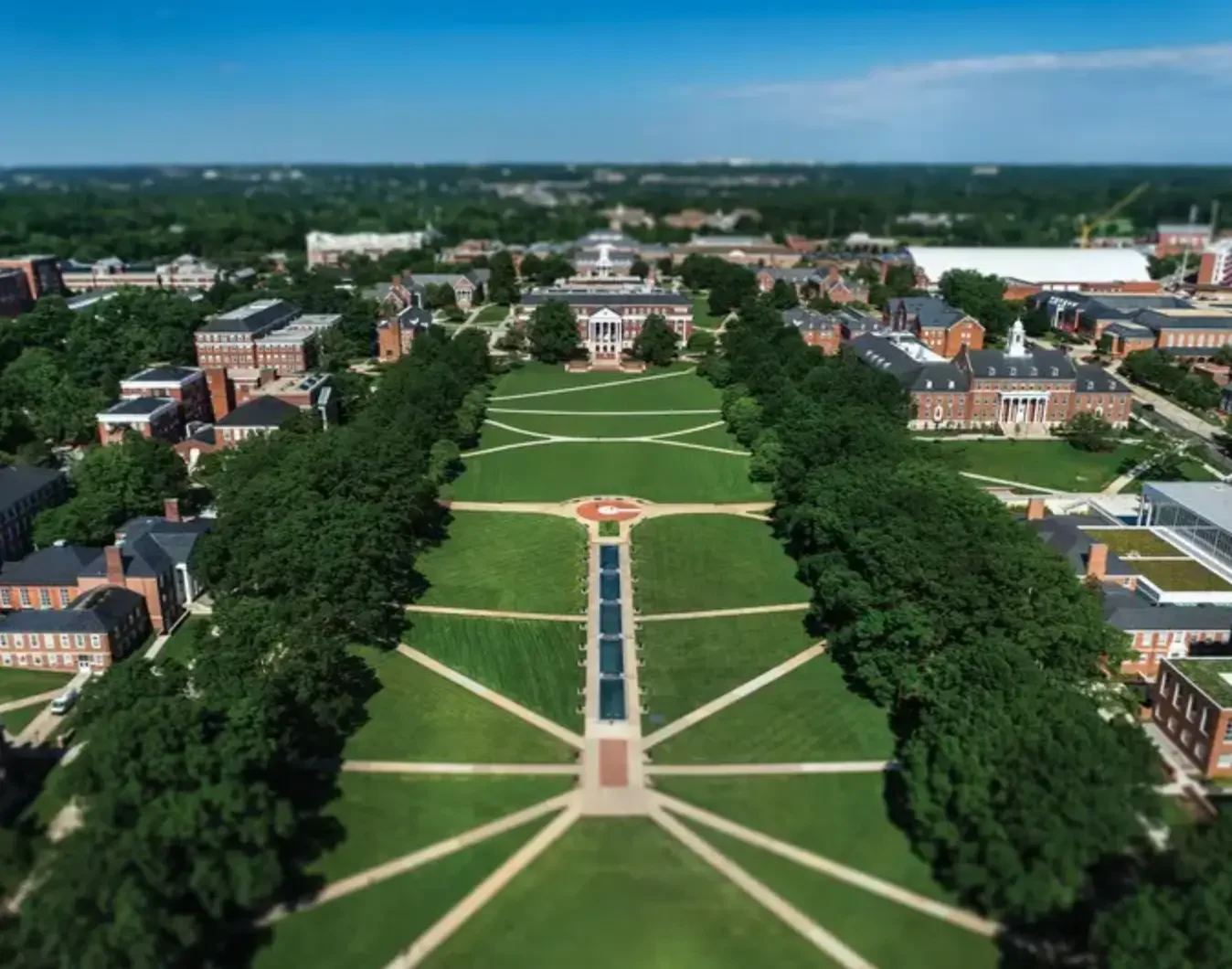Aerial view of a university campus with a large central green lawn, pathways, and multiple academic buildings surrounded by trees.