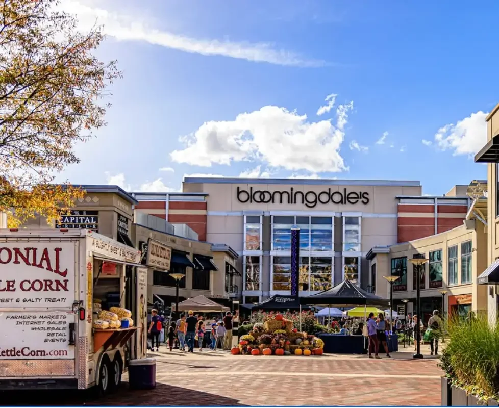Bloomingdale's store with outdoor market stalls and shoppers in a shopping plaza on a sunny day.