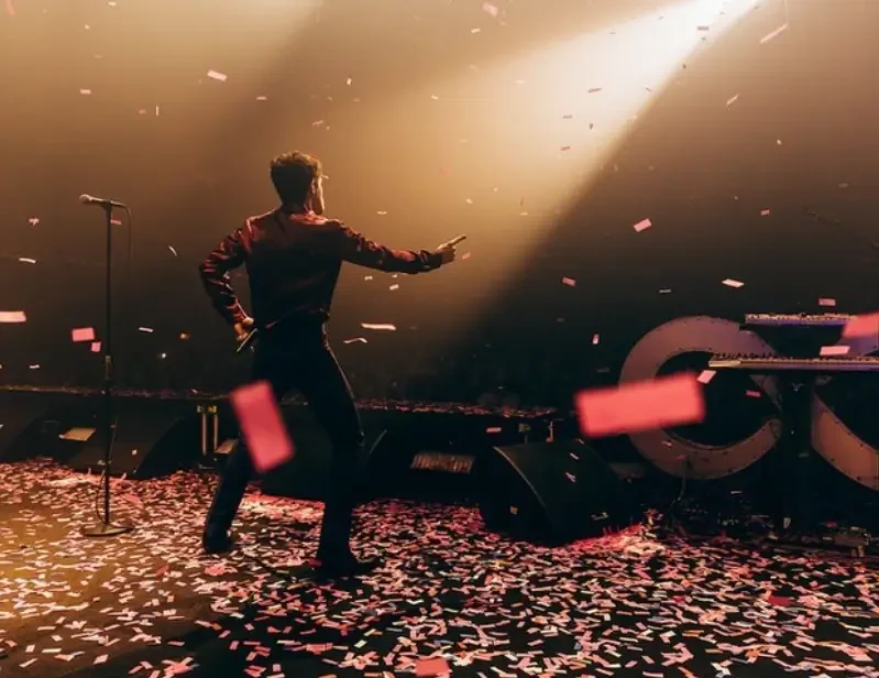 Performer dancing on stage with confetti falling, microphone on stand nearby, illuminated by stage lights, with keyboard in the background.