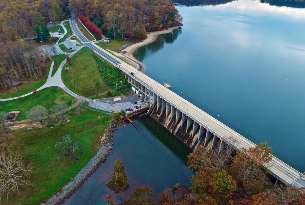 An aerial view of a dam and bridge over a body of water, with surrounding green park and wooded areas.