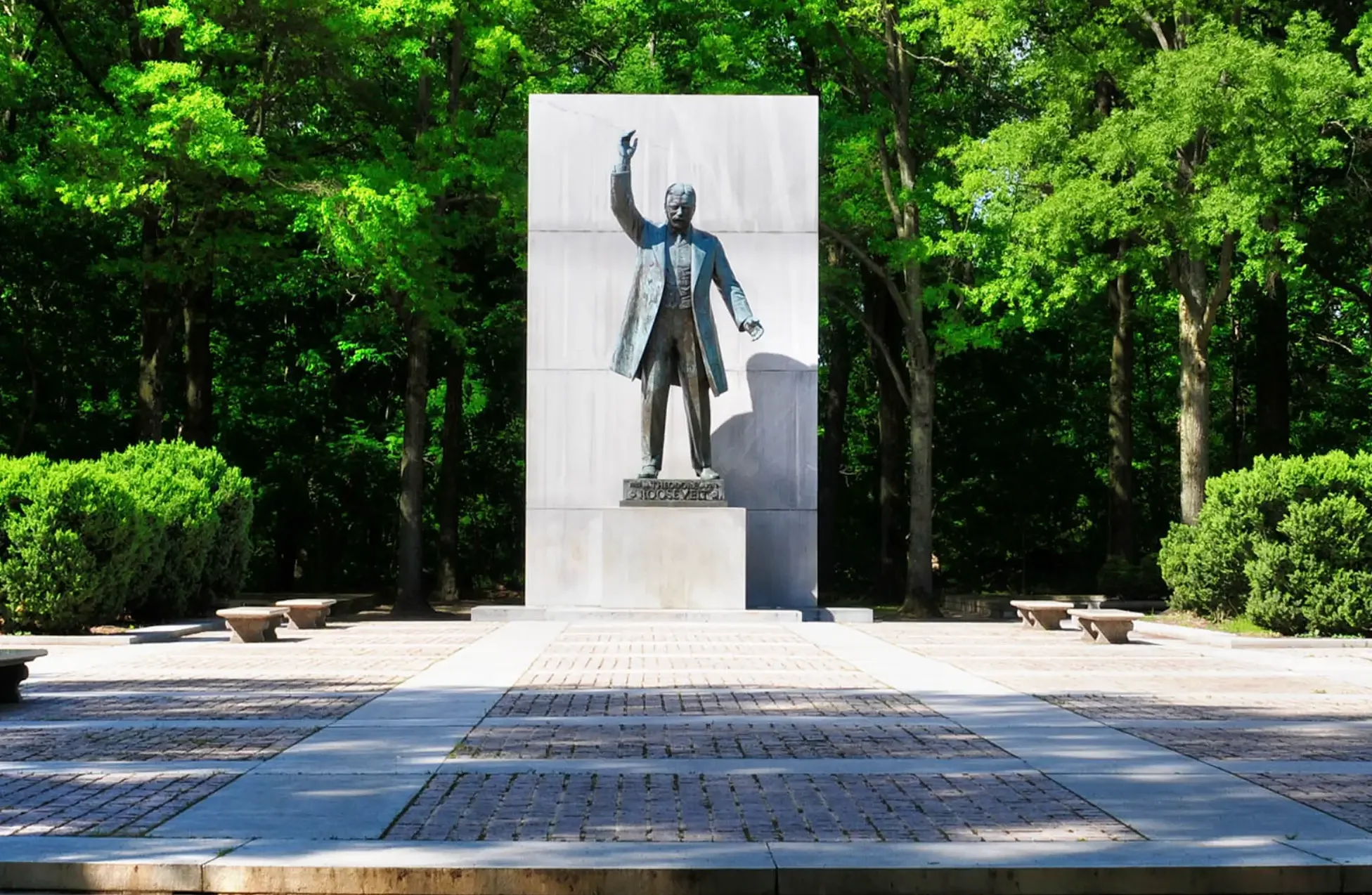 Statue of President Theodore Roosevelt, with one arm raised, located in a park with lush green trees and benches around.