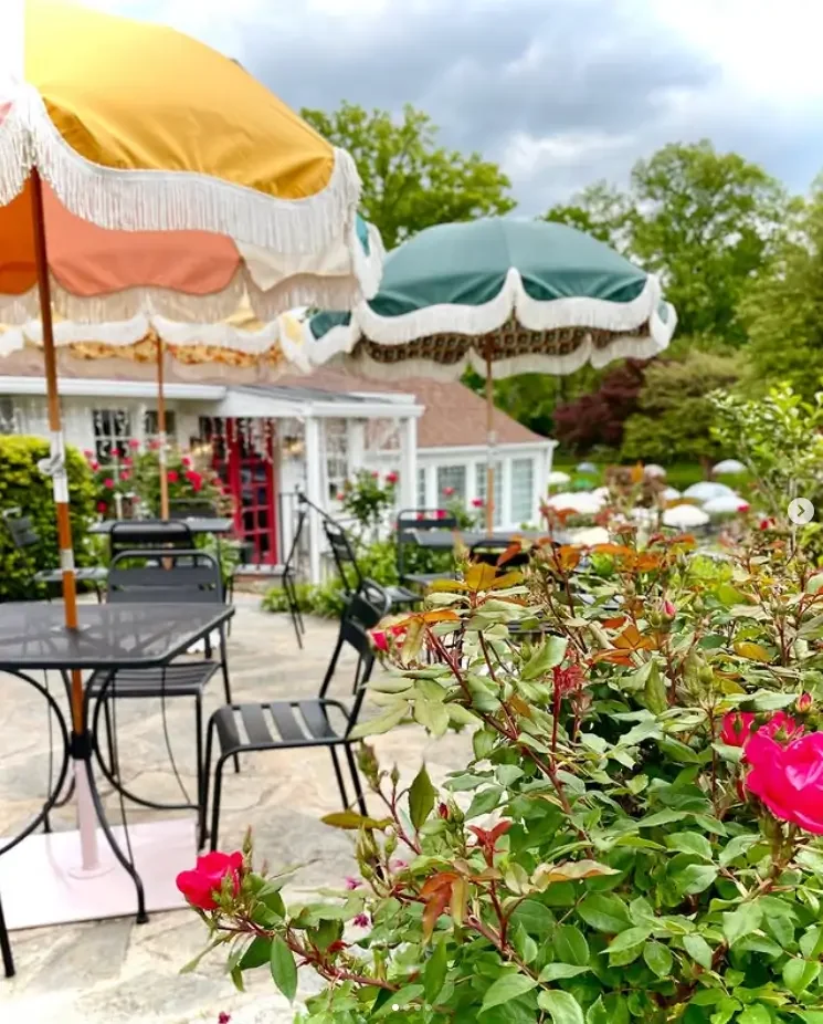 Outdoor patio with black metal tables and chairs, large vintage-style umbrellas, and blooming roses, in a garden setting with a house in the background.