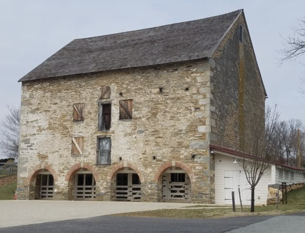 Old stone barn with arched openings, boarded-up windows, and a sloped roof, with trees and a gravel path in the foreground.