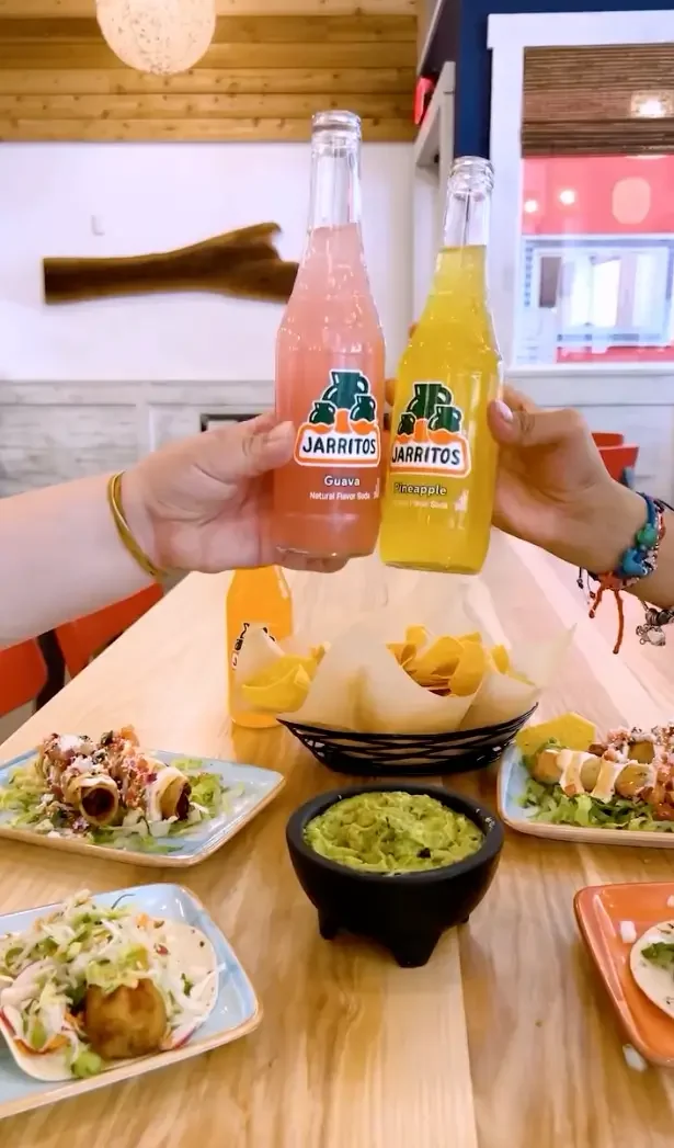 Two people holding bottles of Jarritos soda, one guava and one pineapple, over a table with Mexican food including tacos, chips, guacamole, and tostadas in a restaurant.