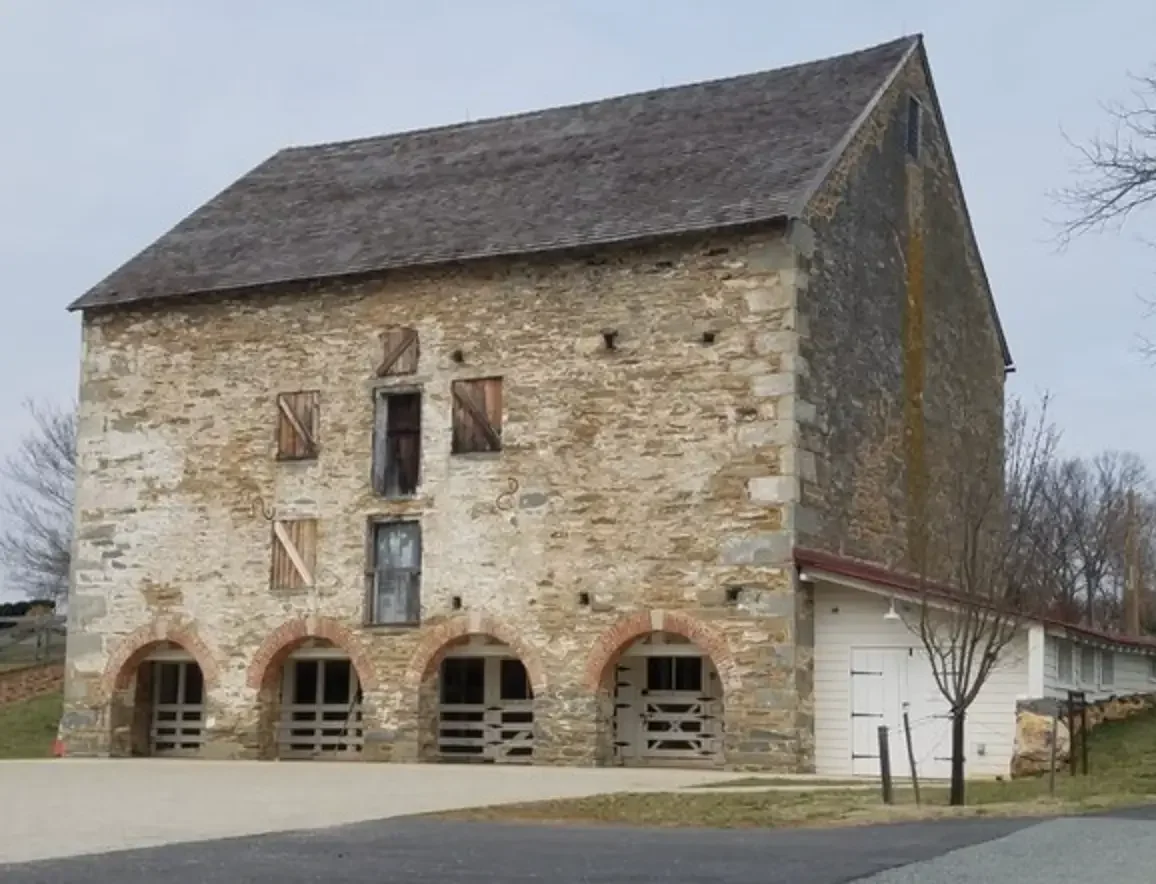 Old stone barn with arched openings, boarded-up windows, and a sloped roof, with trees and a gravel path in the foreground.