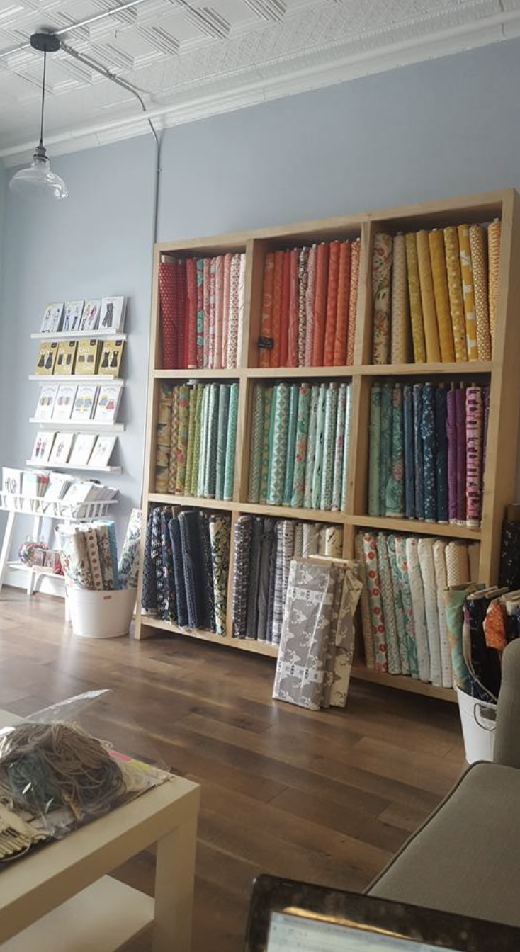 Display of colorful fabric bolts on a wooden shelf in a fabric store, with white books and other retail items around.