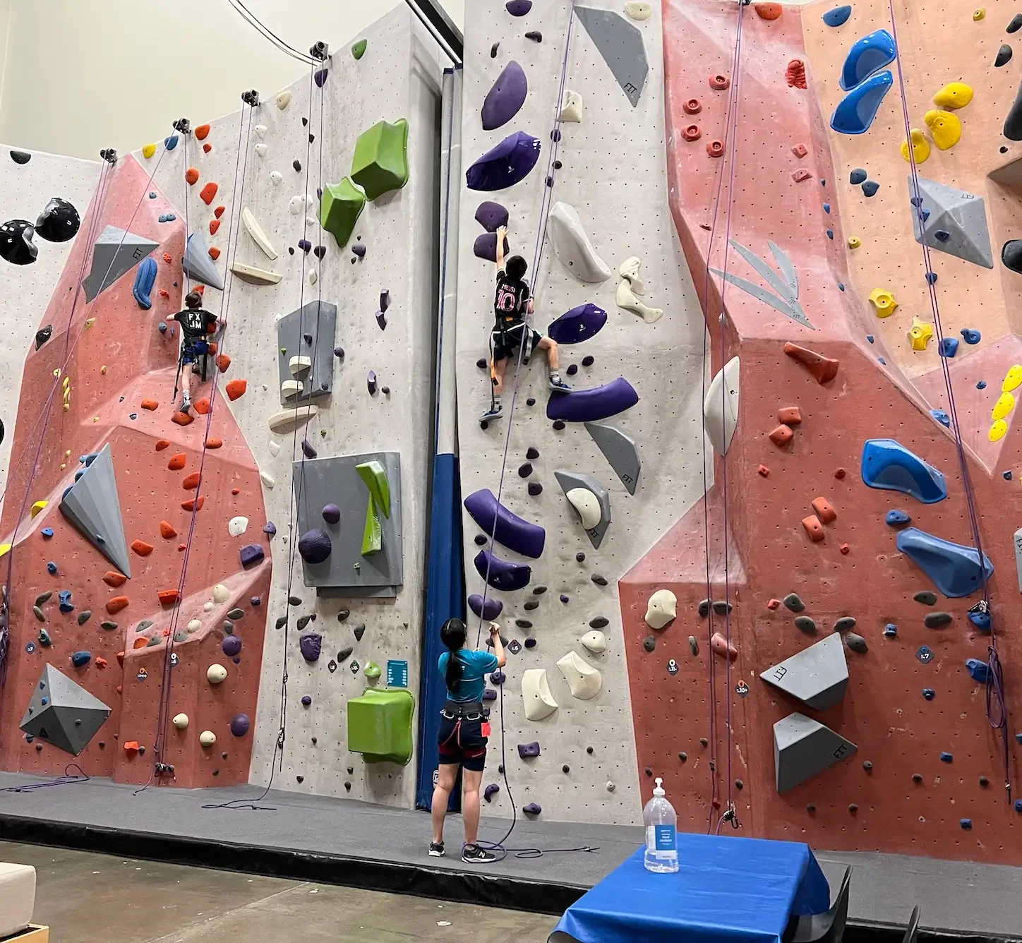 Indoor rock climbing gym with colorful climbing walls and people climbing with harnesses. A woman is belaying at the bottom, watching two children climbing.